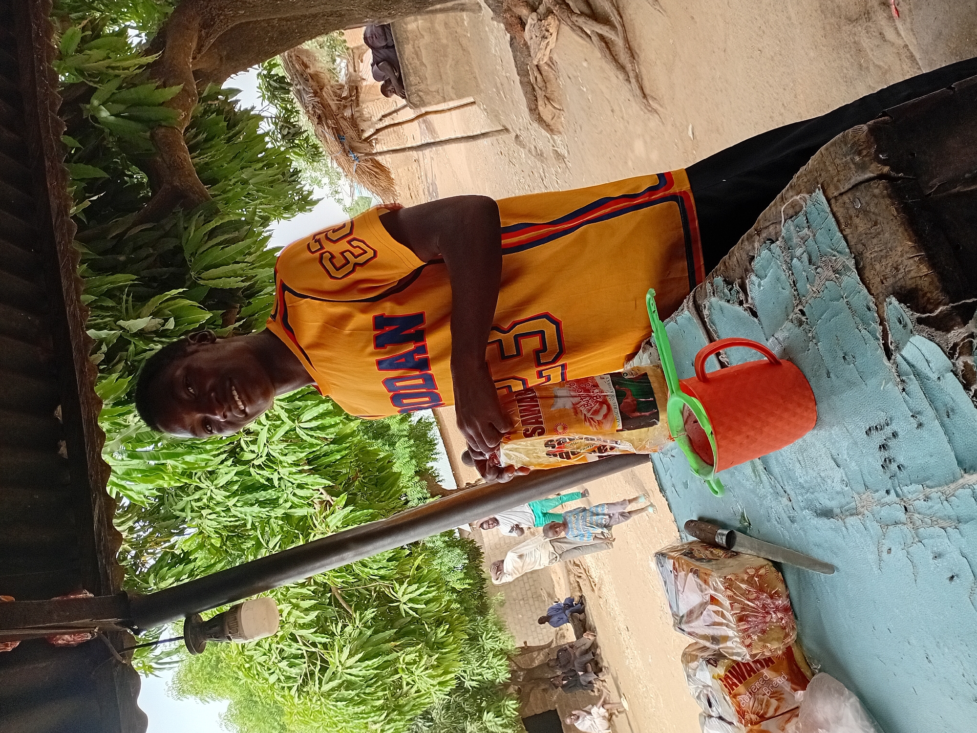 A man wearing a yellow jersey with "JORDAN 23" on it stands at a table outside a structure, likely a small shop, in Nata-Ala, Nigeria. He is smiling and holding a package of "SAWARI Buttered Bread." On the table is a red cup, a green scoop, a knife, and more packaged bread. Other people are visible in the background, standing near a brick structure and under a tree. Trees and foliage are prevalent in the surrounding environment.