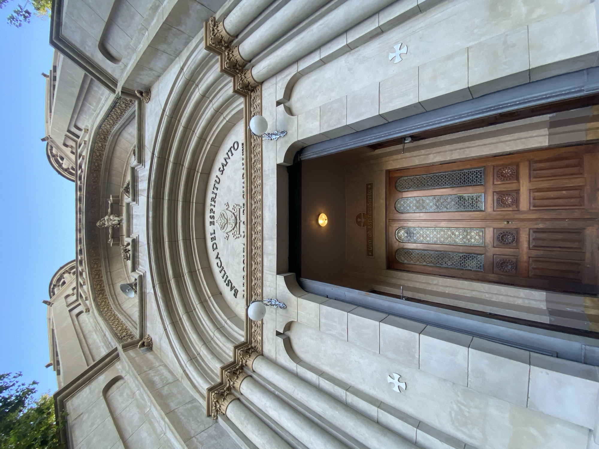 The image depicts the entrance of the Basílica del Espíritu Santo, located in Buenos Aires, Argentina. The camera is positioned looking upwards at the building's facade, capturing the arched entrance and open wooden doors. Architectural details include stone columns with decorative capitals, ornate carvings, and inscriptions reading "BASILICA DEL ESPIRITU SANTO." Two white spherical lamps flank the entrance. Small crosses are embedded in the exterior wall. The doors have decorative panels and glass inserts. A light is visible inside the entrance. The sky is blue in the upper left corner of the image.