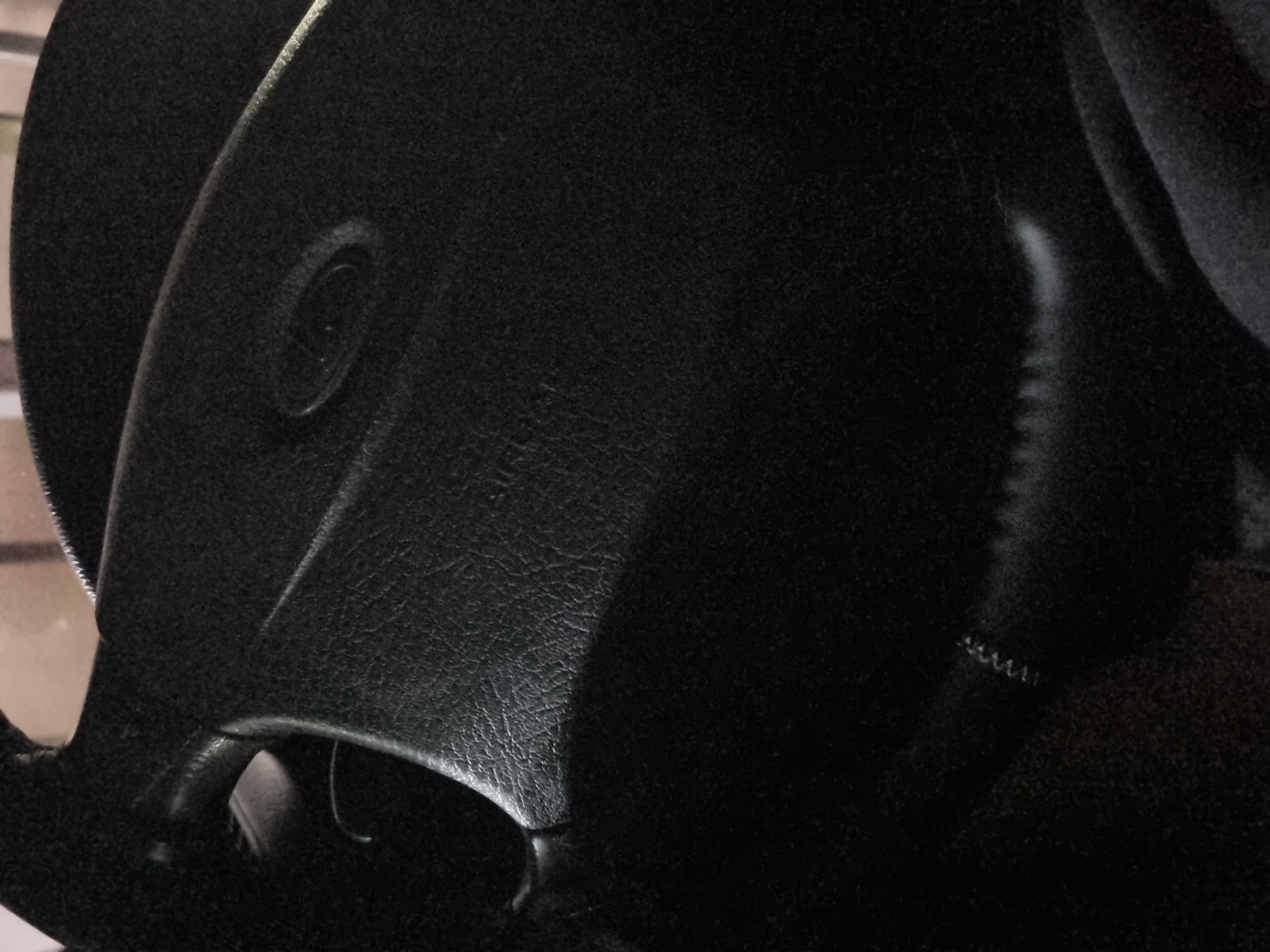 The image shows a close-up view of a black car steering wheel. The steering wheel is made of textured material, likely leather, and has a central airbag cover with a circular embossed logo. A shadow is cast across the lower portion of the steering wheel. The background is dark and out of focus, suggesting it is inside a vehicle. There is no visible indication of the city of Novospasskoye, Russia in the image.