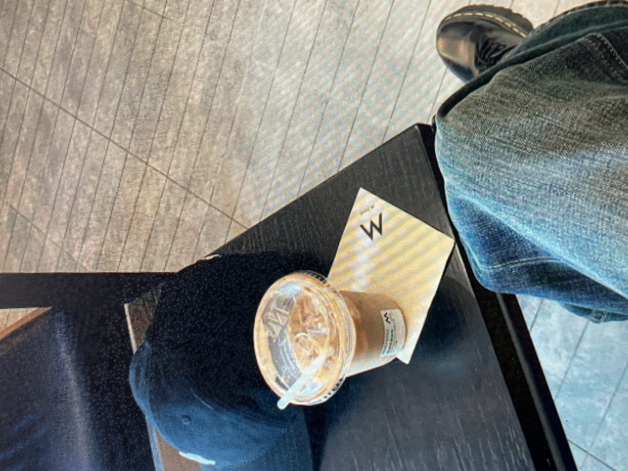 The image depicts a close-up, high-angle view of a black baseball cap resting on a dark-colored surface, likely a table. Adjacent to the cap is a clear plastic cup filled with an iced beverage, possibly coffee, with a white lid and straw. Underneath the cup is a small, folded paper item with a stylized "M" logo printed on it. In the foreground, a person's legs are visible, clad in blue denim jeans and dark-colored shoes with laces, suggesting they are seated. The background shows a tiled floor and what appears to be the side of a wooden bench or chair. The lighting suggests an indoor setting with natural light contributing to the scene. There are no discernible landmarks or contextual clues to indicate a specific location in Sejahtera, Indonesia.