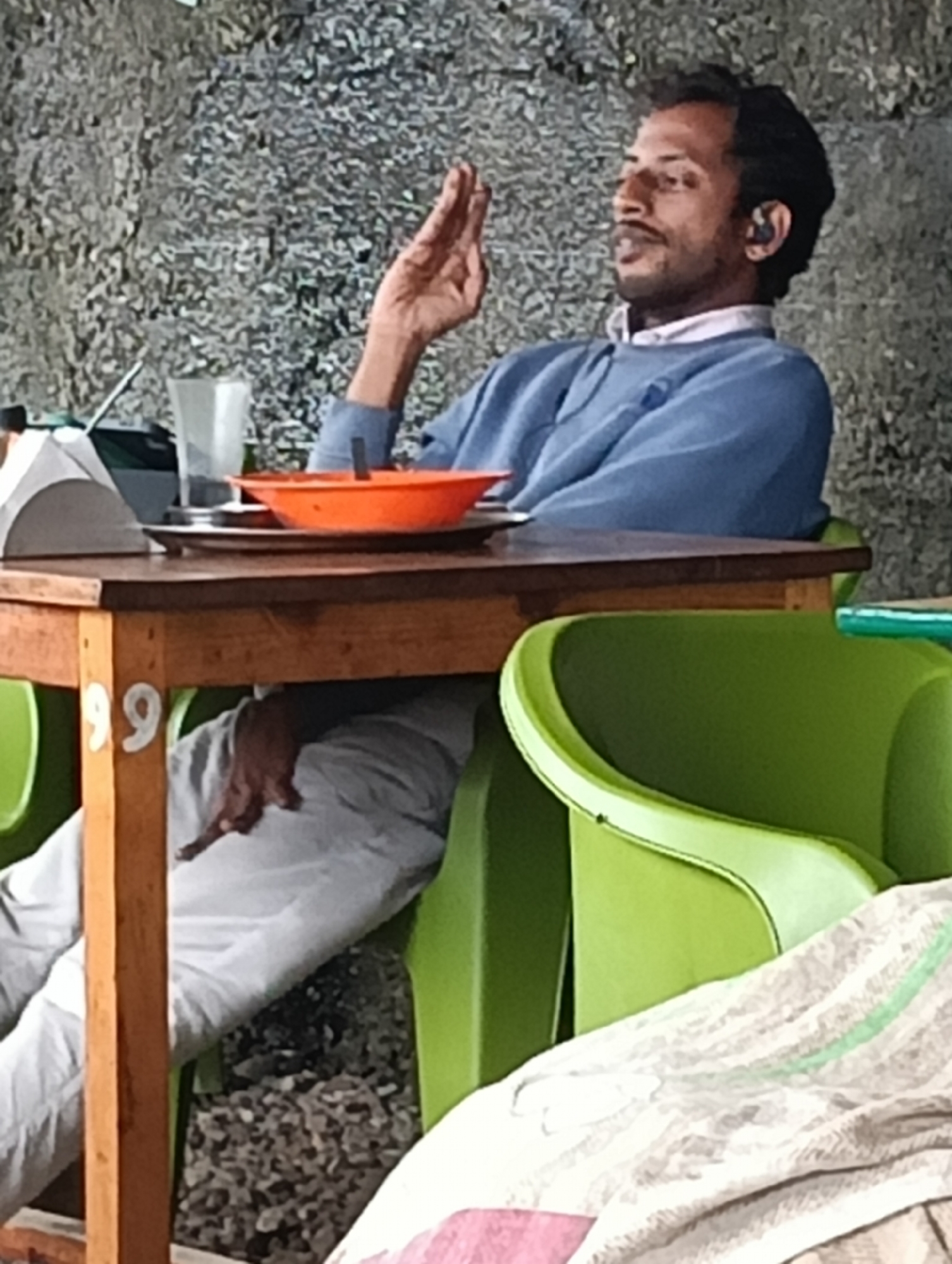 A man sits at a wooden table in what appears to be an outdoor cafe in Haini, India. He is wearing a blue sweater over a white shirt and has a small earbud in his right ear. His left hand is raised, with his fingers extended. In front of him on the table are an orange bowl, a plate, and a glass of water. A white tent-like object is also visible on the table. The man is seated in a green plastic chair. The background is a textured, gray wall. The legs of the table are visible, with the number "99" painted on one of them. Another green plastic chair is visible to the right of the table. Under the table, a person's legs, clad in light-colored trousers, are visible, resting on a light-colored fabric. The ground beneath the table is covered in small stones.