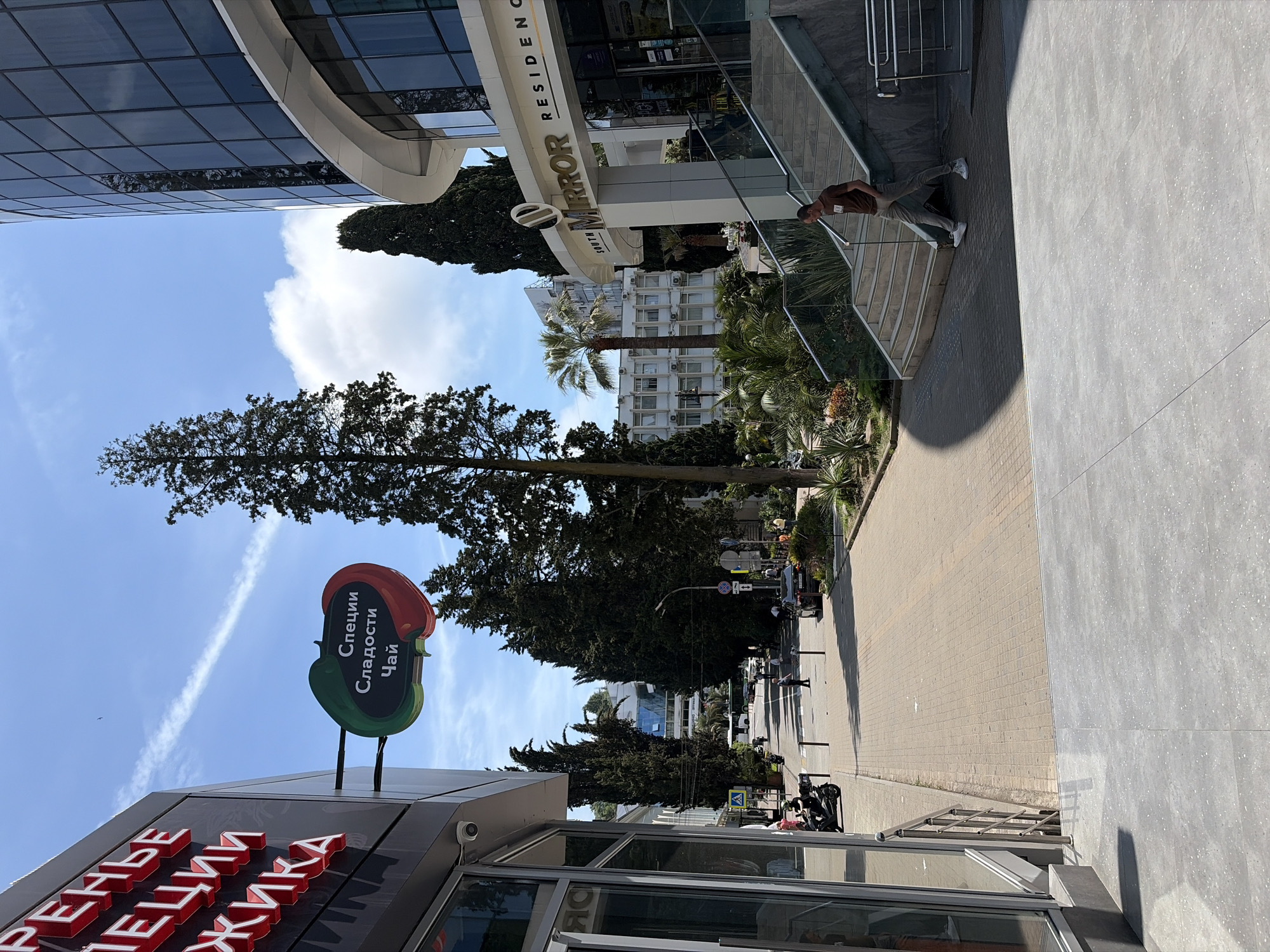 This detailed image captures a sunlit urban street scene in Sochi, Russia, presenting a mix of modern and traditional architecture with lush subtropical vegetation under a clear blue sky.
In the immediate foreground, a light grey tiled ground surface spans the width of the frame. To the left, a portion of a dark-paneled building facade is visible, featuring large, red, three-dimensional Cyrillic lettering, partially reading "РЕНЬЕ", "ПЕЦИИ", and "ЖИКА". A small security camera is mounted on this facade. Extending from this building, a distinctive horizontal sign shaped like two chili peppers (green on the left, red on the right) displays white Cyrillic text: "Специи" (Spices), "Сладости" (Sweets), and "Чай" (Tea).
To the right, a multi-story, modern building with a curved, dark glass facade is prominent. Above its main entrance, a prominent sign reads "SOUTH MIRROR RESIDENCE". A wide, multi-tiered staircase made of light-colored concrete, featuring glass and metal railings, descends from this building's entrance. A male individual, wearing a brown t-shirt, light-colored trousers, and white sneakers, is depicted mid-stride descending these stairs, looking down towards his feet. Lush green landscaping, including a tall palm tree and various shrubs and succulents, surrounds the base of this building and flanks the staircase.
The central area of the image reveals a pedestrianized street or walkway paved with light-brown interlocking pavers, extending into the background. Black bollards are intermittently placed along the sides of this path. A black motor scooter is parked on the left side of the street, near the dark building. Further down the street, several pedestrians are visible, along with parked vehicles and a white public transit bus in the mid-distance. Streetlights and various traffic signs are also present.
Numerous tall, dark green coniferous trees, likely cypress, line the street and are interspersed among the buildings, providing significant vertical elements. Additional palm trees are scattered throughout the scene, particularly near the "Mirror Residence" and in the background.
The background features a cluster of buildings showcasing different architectural styles, including a multi-story white building with many windows and visible exterior air conditioning units. The sky is bright blue with scattered white cumulus clouds, and a prominent white aircraft contrail stretches diagonally across the upper left quadrant. The scene is brightly illuminated by direct sunlight, casting distinct shadows.