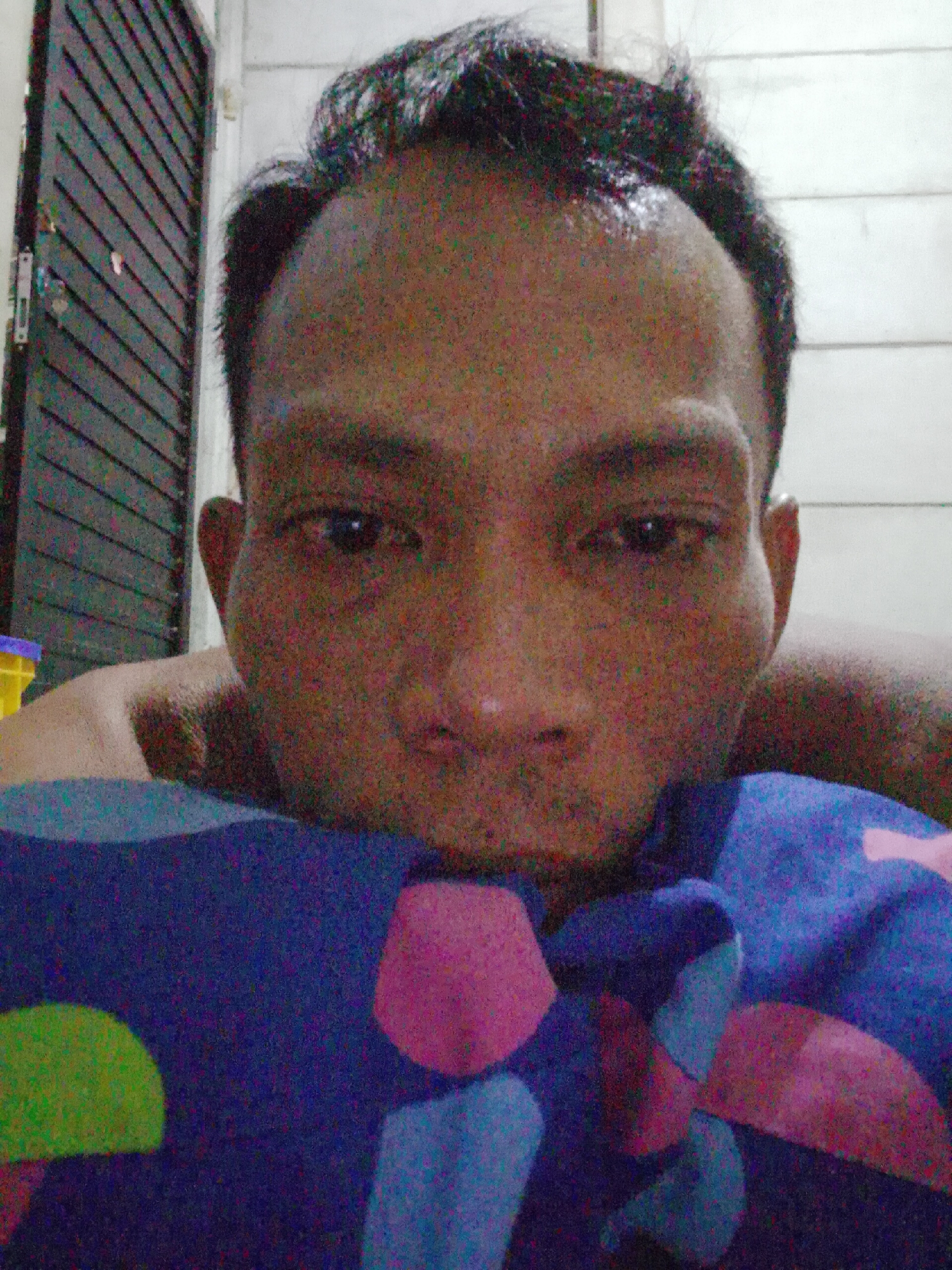 An adult man is looking directly at the camera, possibly from a low angle or lying down. His head is close to the camera. He has short, dark hair and appears to be wearing or lying under a blanket or sheet with a pattern of irregular shapes in colors of blue, pink, and green. Behind him, on the left side of the frame, is a dark, shuttered or slatted door, and on the right is a white wall. Based on the prompt, the location is potentially in Bukit Ketok, Indonesia.