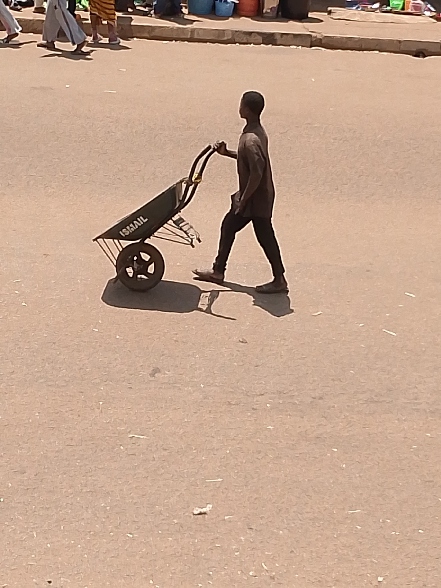 A young Black male walks down a paved road in Bukuru, Nigeria, pushing a dark-colored wheelbarrow. He is wearing a dark brown short-sleeved t-shirt, dark pants, and brown shoes. The wheelbarrow has a black metal frame and a black tire with a multi-spoke design. The word "ISMAIL" is visible in white lettering on the side of the wheelbarrow's basin. The ground is a sandy, light brown asphalt. In the background, the blurred forms of other people, some wearing light-colored clothing and others patterned garments, are visible along the side of the road, suggesting a marketplace or busy street. Partially obscured by the people and wheelbarrow are some cylindrical blue objects, possibly buckets or containers. The lighting suggests a bright, sunny day, casting distinct shadows.