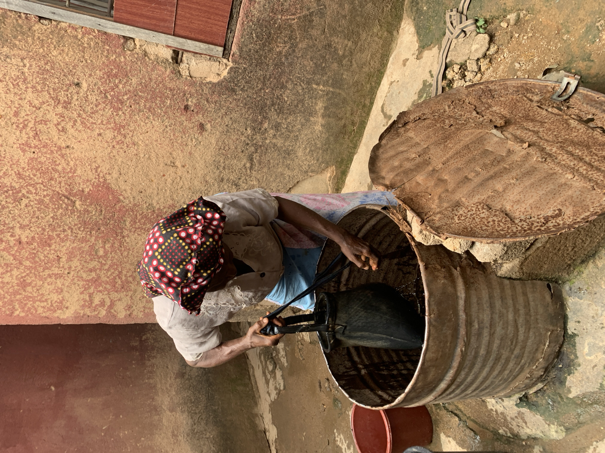 A woman wearing a red and black patterned headscarf, a light-colored shirt, and light blue shorts is drawing water from a well located outdoors, adjacent to a reddish-brown wall with a small window. The well is constructed from a large, rusted metal barrel with its lid open and resting nearby. She is using a black bucket with black rope handles to draw the water. A red bucket is located near the base of the well. The location is likely Jos, Nigeria.