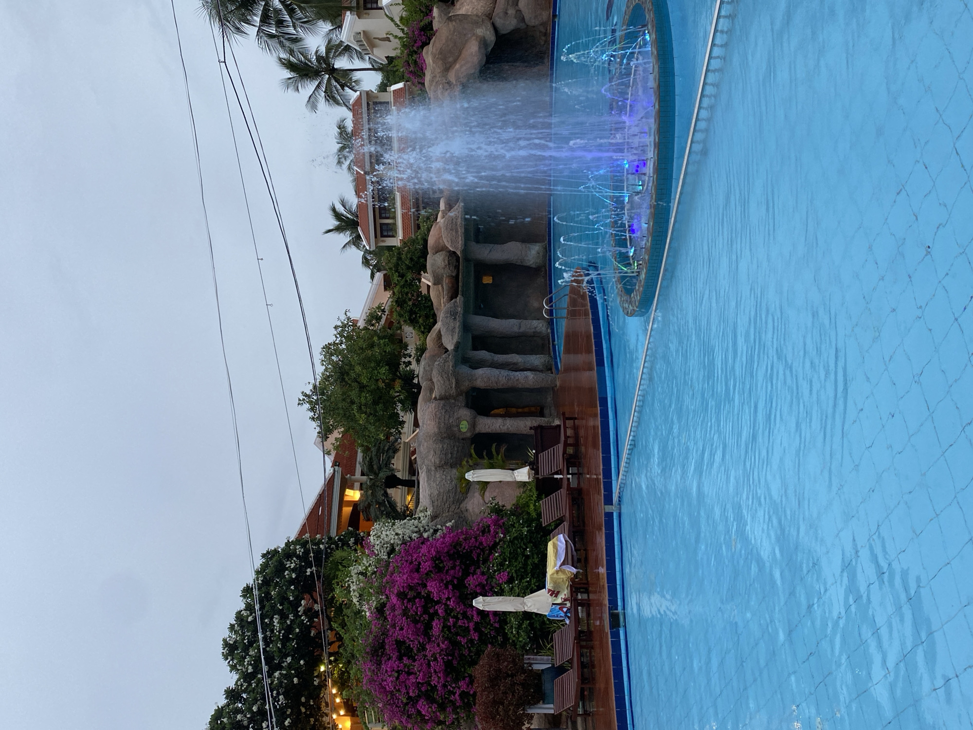 The image shows a swimming pool with a fountain in Phu Thuy Ward, Vietnam. The pool is rectangular with light blue tiles. The fountain, located on one side of the pool, features a central geyser and a ring of water jets illuminated with blue lights. Faux rock formations and lush vegetation surround the fountain. Lounge chairs and closed umbrellas are positioned near the pool edge. Buildings with tiled roofs are visible in the background among the trees. Power lines stretch across the sky. The weather appears overcast.