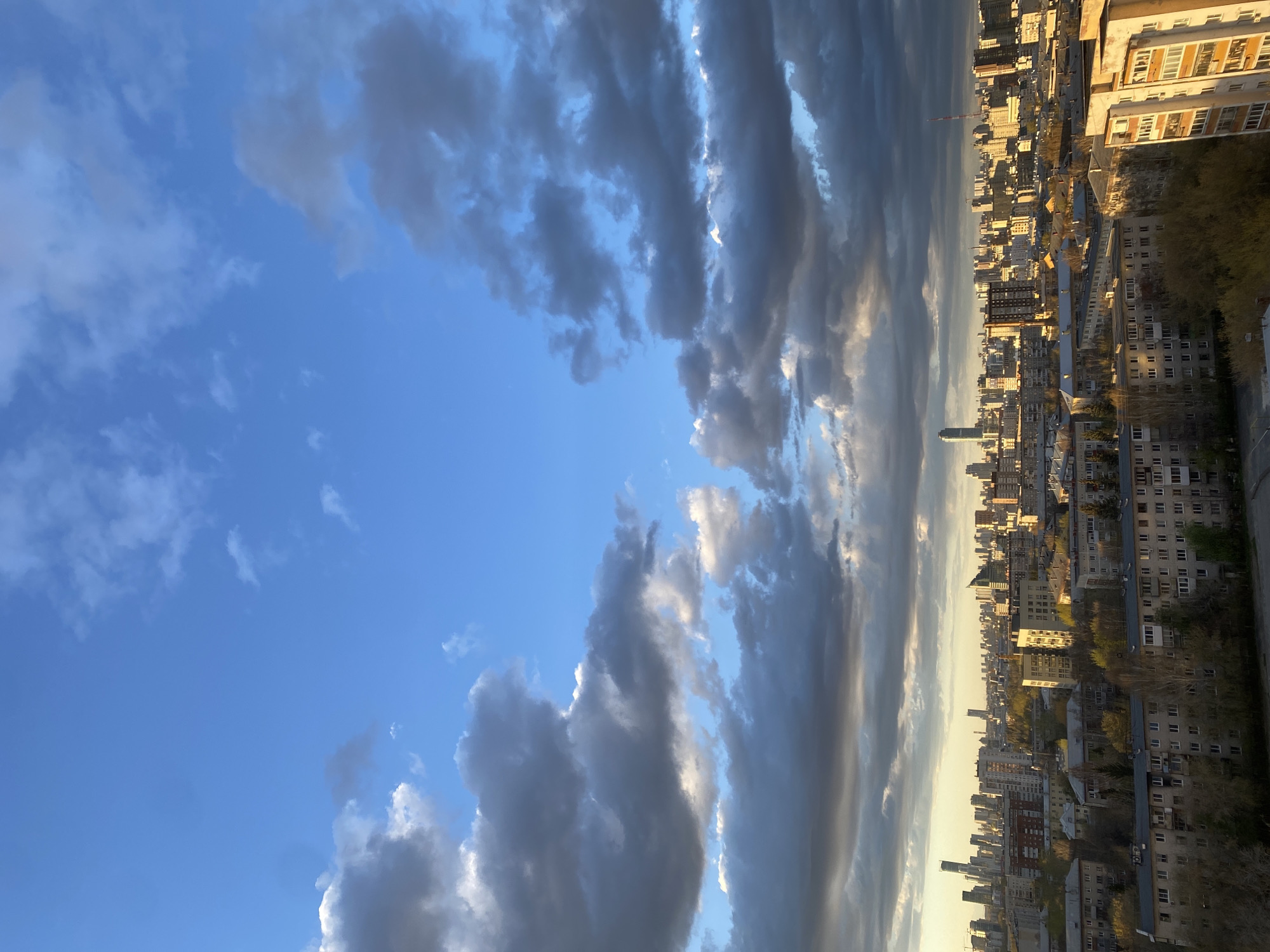 This image presents an expansive urban landscape viewed from a high vantage point, with a dramatic sky overhead. The city stretches across the lower portion of the frame, bathed in the warm, golden light of what appears to be late afternoon or early evening.
The sky is a prominent feature, displaying a striking contrast. On the left, it is a clear, vibrant blue with scattered, wispy clouds. Towards the center and right, large, dark, and heavily textured clouds dominate, their edges brightly illuminated by the low sun, creating a sense of depth and movement. Patches of brighter sky are visible between these denser cloud formations.
Below the dynamic sky, the cityscape unfolds. Numerous buildings of varying heights and architectural styles populate the urban sprawl. In the foreground, older, multi-story residential buildings with multiple windows are visible. Further into the distance, a dense cluster of modern high-rises and skyscrapers rise, with their facades catching the warm sunlight. The sun's low angle casts long shadows across the city, highlighting its contours and density. Hints of green foliage from trees are interspersed among the buildings, suggesting urban parks or street-side greenery.
No people or discernible text are visible in the image. The scene captures the vastness of an urban environment under a beautiful and imposing sky, likely at sunset or sunrise, creating a serene yet grand atmosphere. Given the context, this is a view of Yekaterinburg, Russia.