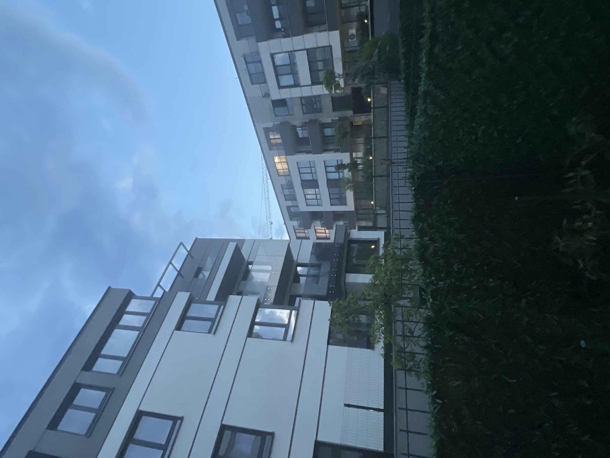 The image depicts an exterior view of modern apartment buildings against a twilight sky in an unspecified urban location. The buildings are multi-story structures with a contemporary architectural design, featuring large windows and balconies. The foreground is dominated by a dense green hedge and a black metal fence. In the background, another apartment building is visible, partially obscured by foliage, with some windows illuminated, suggesting interior lighting. A construction crane is visible in the distance, extending above the buildings. The sky is a gradient of deep blue to lighter blue, with some wispy clouds. The overall scene conveys a sense of urban development and residential living at dusk.