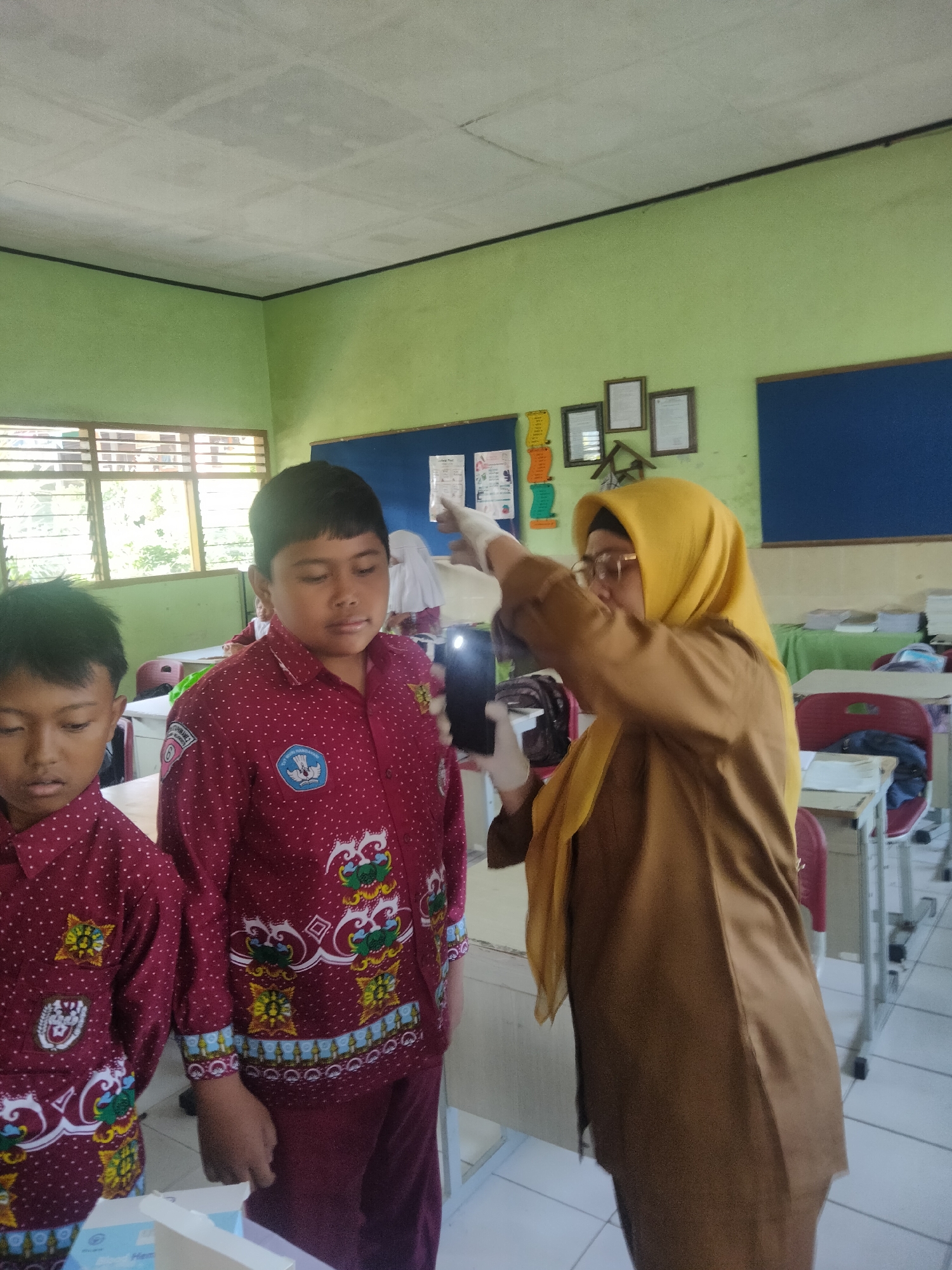 In a classroom setting, a woman wearing a yellow hijab and a brown tunic, with white gloves on her hands, appears to be taking a photo of a young boy. The boy, who is wearing a maroon uniform shirt with white polka dots and a school emblem, stands facing forward. Another young boy, also in a similar maroon uniform, is partially visible to the left. The classroom has light green walls, a white tiled floor, and rows of desks. A window with slatted blinds is visible on the left, and framed pictures and a bulletin board are on the back wall. The scene suggests an activity or event taking place in a school in Tretes, Indonesia.