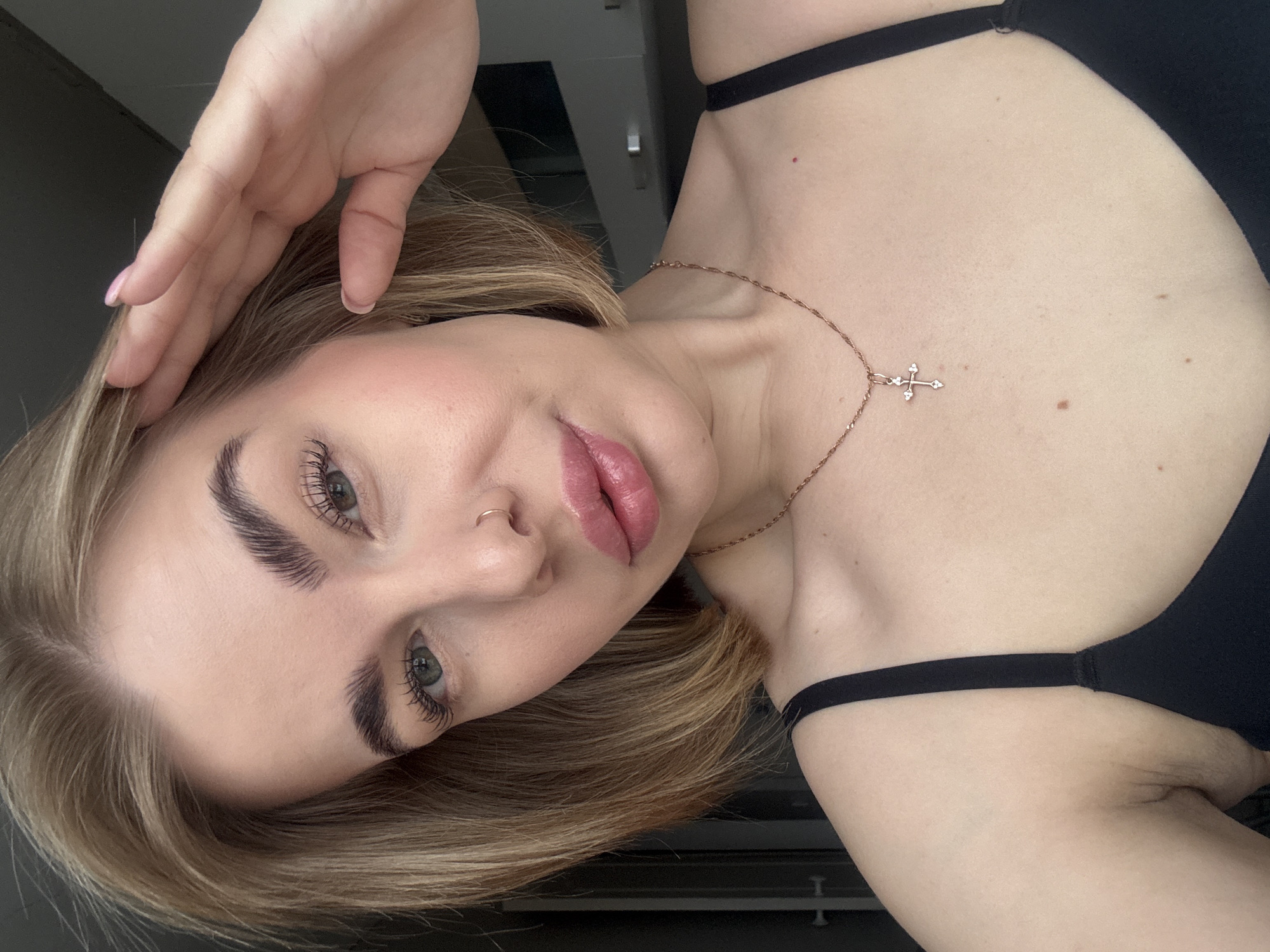 A woman with blonde hair poses for a selfie-style photo, resting her hand on her head. She is wearing a black top and a delicate gold necklace with a cross pendant. She has full lips, fair skin, and prominent eyebrows. A small ring is visible in her left nostril. The background appears to be an interior space, possibly a bedroom, with a cabinet or wardrobe partially visible. The location is Chaikovsky, Russia.