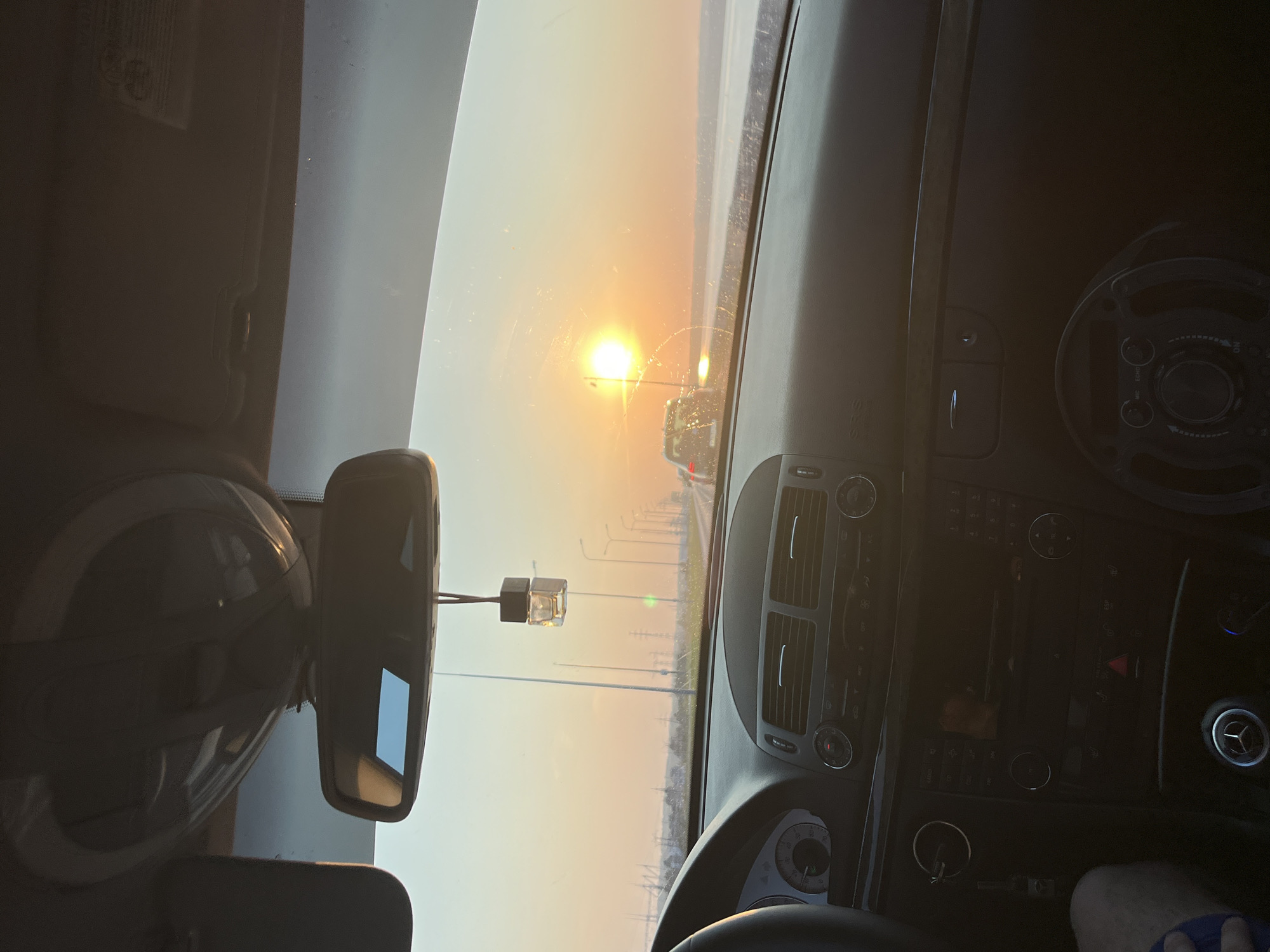 The image is an interior view from the passenger seat of a car, facing forward through the windshield. The car is on a road with streetlights and other vehicles visible in the distance. The sun is low in the sky, casting a warm, orange glow and creating a bright glare on the windshield. The dashboard of the car is visible, with the steering wheel on the left side. A car air freshener hangs from the rearview mirror. The background shows a flat landscape with some structures and what appears to be a body of water. The setting suggests it is either sunrise or sunset. The location is identified as Chaikovsky, Russia.