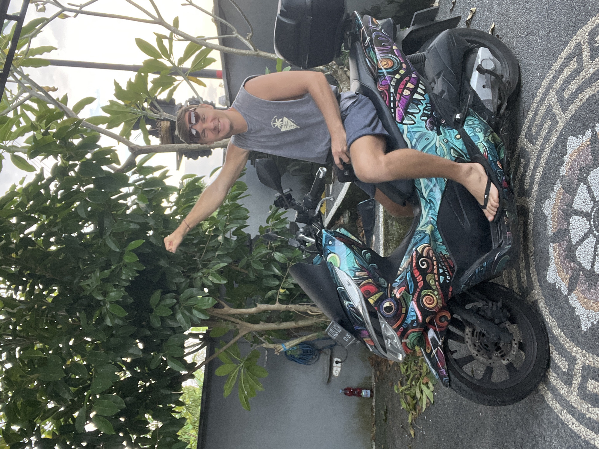 The image captures a man sitting on a vividly decorated motorcycle. He's wearing a grey tank top, blue shorts, sunglasses, and flip-flops. He raises his left arm with a clenched fist. The motorcycle, displaying various colorful patterns, is parked against a grey wall. A tree stands to the left of the wall. The surrounding area is outdoors on what appears to be a patterned stone path. Based on the location in the city of Sukawati, Indonesia, the environment suggests a tropical or subtropical setting. A water bottle is on the ground near the motorcycle.