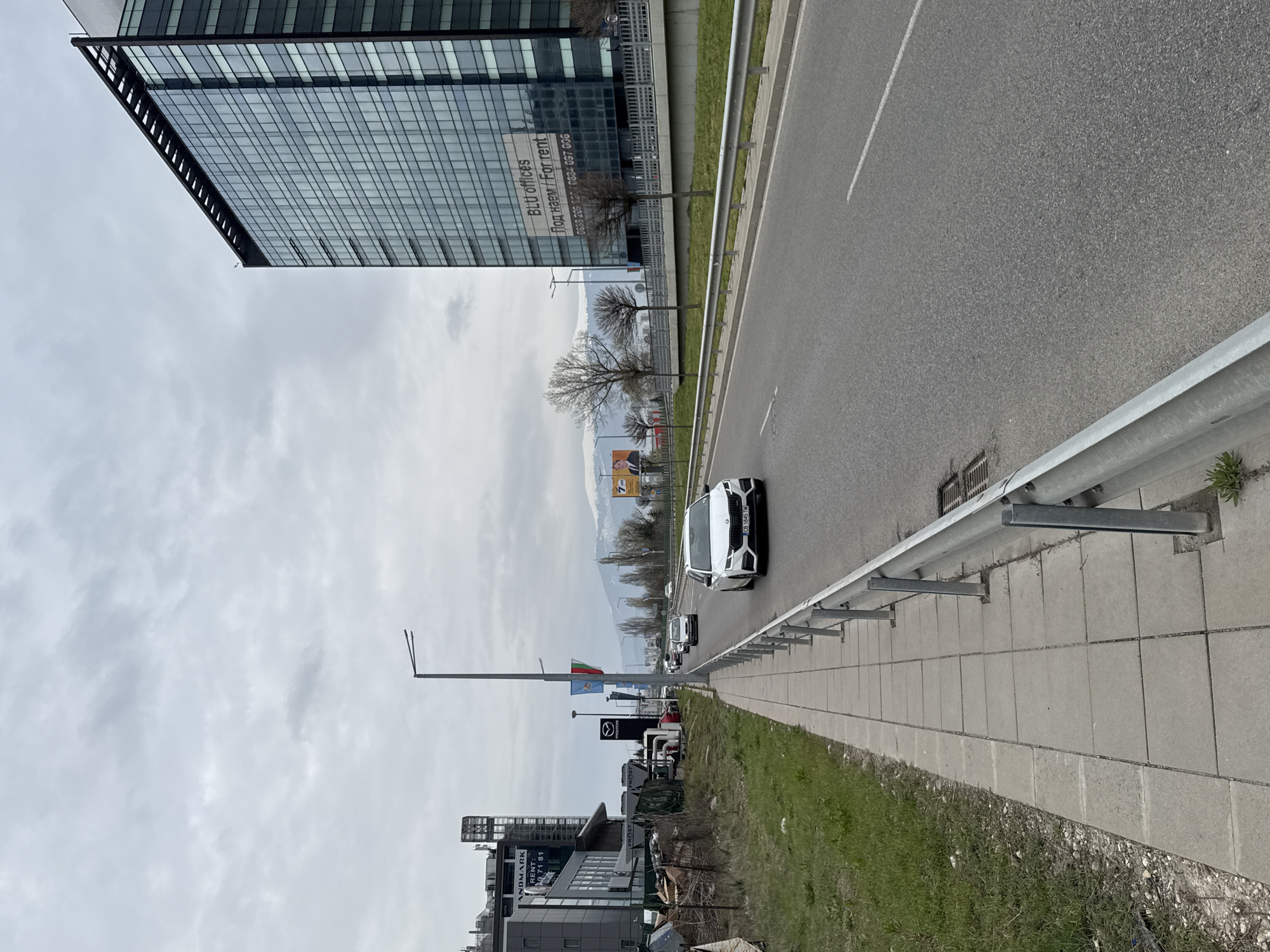 The image shows a street scene in Sofia, Bulgaria. A modern glass building labeled "BLU offices" with rental information is visible on the upper left. A street runs alongside the building, separated by a grass strip and a metal guardrail. Cars are traveling on the road, including a white car in the foreground with the license plate "CB 1845 TM". Further down the street, there are signs for Mazda and Frost. In the background, mountains are visible under a cloudy sky. There are buildings on both sides of the street. The foreground shows a concrete sidewalk and a grassy verge.