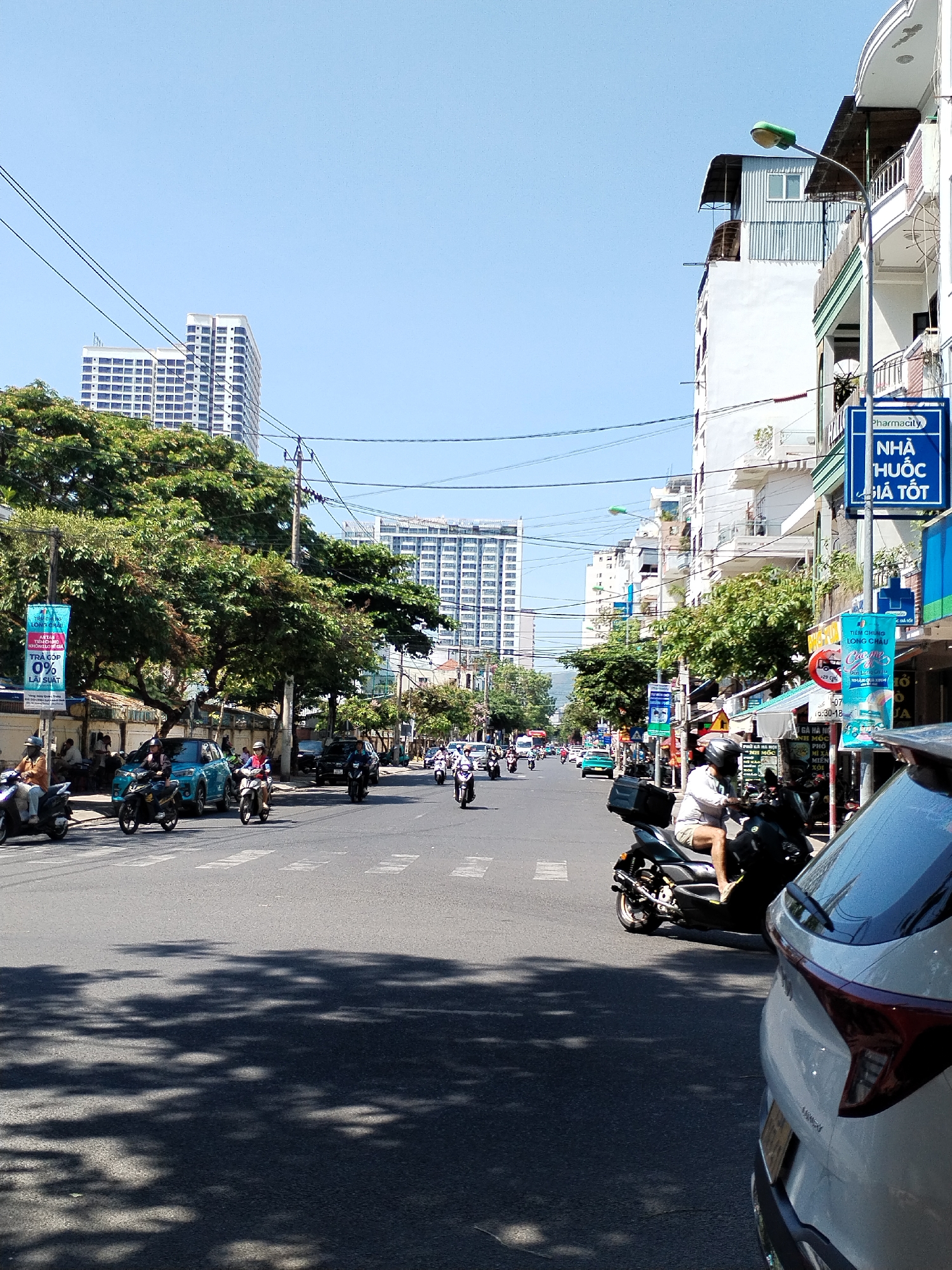 The image is an eye-level view of a street scene in Nha Trang Ward, Vietnam, captured on a sunny day. The asphalt road, patterned with shadows, stretches into the distance, where various buildings can be seen. On the left, tall buildings rise above the trees lining the street. Power lines crisscross overhead. Motorbikes populate the road, some in motion, others stationary at the side. Banners and street signs are visible on the right side of the street. A white car occupies the right corner of the frame, partially obscuring the view.