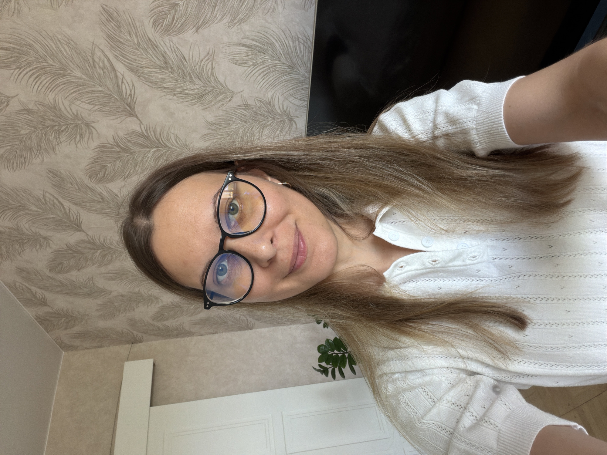 A woman with long, blonde hair and glasses is taking a selfie indoors. She is wearing a white, short-sleeved shirt with a collared neckline and buttons. The background appears to be a room with patterned wallpaper and a white door on the left. A potted plant is visible to the left of the woman. A dark object, possibly a television, is present in the background to the right. The lighting suggests it is daytime. The image does not contain any information that can be used to determine the specific location within Izhevsk, Russia.