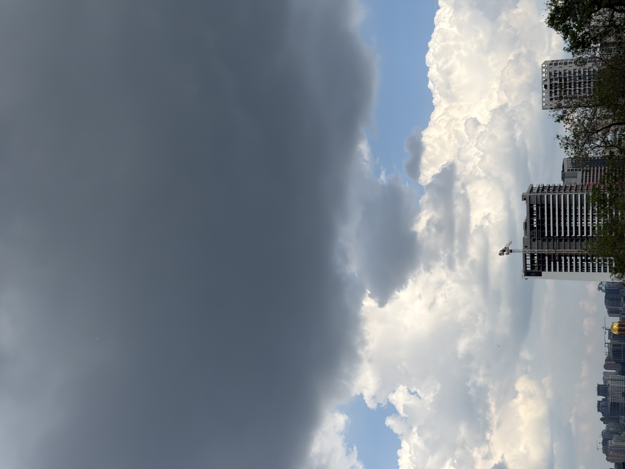 The image displays a dramatic and sharp contrast in the sky, vertically divided into two distinct weather patterns. The left half of the frame is dominated by a mass of dark, heavy, ominous gray clouds, suggesting an approaching storm or heavy rain. In stark contrast, the right half of the image features bright, voluminous cumulus clouds against a clear blue sky, indicating fair weather.
Beneath the brighter clouds on the right, a city skyline is visible. Several modern high-rise buildings, some appearing to be under construction with exposed framework and a visible tower crane, stand tall. Patches of green foliage from trees are seen on the far right, partially framing the buildings. In the distant cityscape, a distinctive golden dome of a building, likely a church, is discernible, serving as a location cue for Kyiv, Ukraine.
The scene captures a dynamic weather event, likely a weather front passing over the city during daytime, given the bright illumination on the fair-weather side. There is no discernible human activity, and no text is visible within the image.