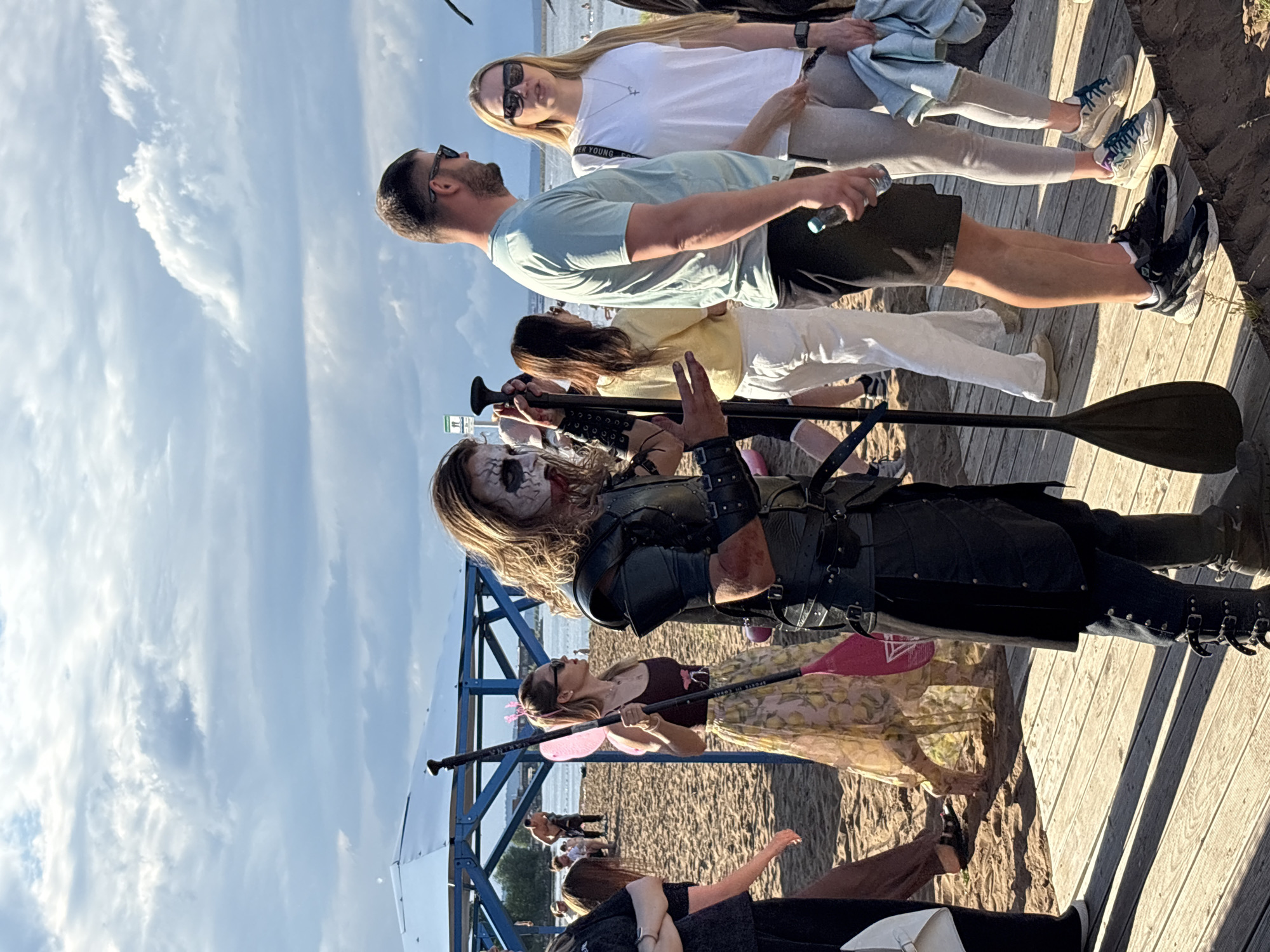 This media file captures a group of people on a wooden boardwalk near a sandy beach, with a body of water and a blue sky with scattered clouds in the background. The scene appears to be set during the daytime, with sunlight casting distinct shadows, suggesting a clear and possibly warm weather day.
The subjects include several individuals. In the foreground, a person in what looks like a costume, with face paint resembling a skeleton or zombie, is holding a paddle. Behind them, a woman with blonde hair and sunglasses is also holding a paddle, wearing a patterned skirt and a dark top. Further back, a man in a light blue t-shirt and dark shorts is looking upwards. Two other women are visible, one in a white t-shirt and grey leggings, and another in a light-colored top and white pants. The overall atmosphere seems casual and perhaps related to an outdoor event or gathering.
There's a small red and white structure with blue metal framing in the middle ground, possibly part of a beach setup or pavilion. The wooden boardwalk is made of weathered planks. The activity appears to be people socializing outdoors, with paddles suggesting a potential connection to water sports like paddleboarding. The image is oriented vertically.