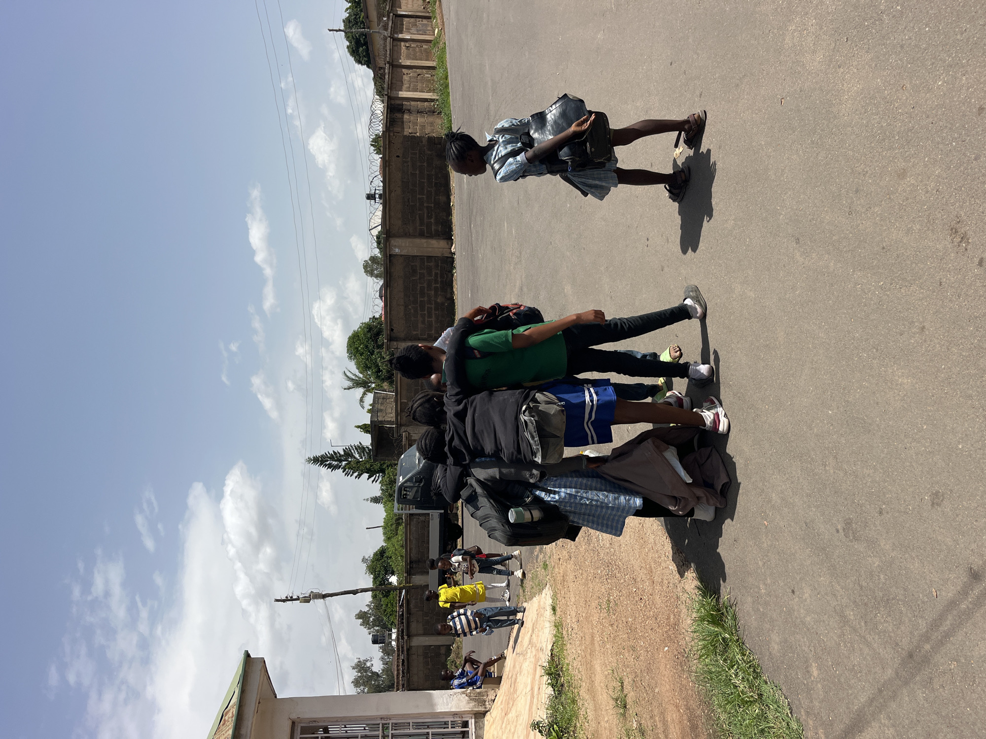 In an outdoor scene in Unknown, Unknown, a group of children stands on a paved street next to a dirt path with grass. A building with a metal roof and a window is visible to the left. A black truck is parked in the background, behind a low brick wall topped with barbed wire, and trees and a utility pole.
In the foreground, a group of five children is clustered together. Two boys have their arms around each other's shoulders. One boy wears a green t-shirt and black pants. His companion wears a black hoodie and blue shorts. A third boy is behind them, wearing a black jacket and plaid skirt. A fourth child, a girl, stands slightly apart to the right, wearing a blue and white plaid dress and a backpack. She is looking to the right. Another child, a girl in a yellow dress, stands further to the left with two other children. One of these children is wearing a blue and white striped shirt and blue pants, and another is wearing blue athletic shorts and is seated on a low step. The children are carrying backpacks and jackets, suggesting they may be students. The sky is partly cloudy with white clouds against a blue sky. The lighting suggests it is daytime, likely late morning or early afternoon.