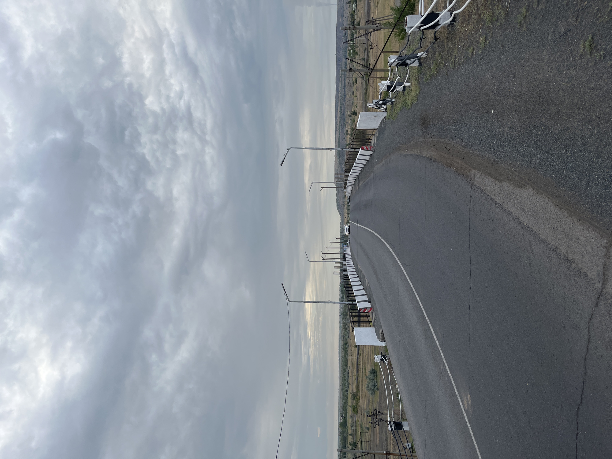 The image shows a two-lane asphalt road curving to the right, likely a bridge, under a cloudy sky. The road is bordered by a white and black striped guardrail on both sides. Several streetlights are visible along the guardrail. In the distance, a flat, dry landscape with sparse vegetation and some industrial structures, including what appears to be a smokestack emitting smoke, is visible. A white car is driving away from the camera on the right lane of the road. The scene appears to be in an industrial or semi-rural area. This view is consistent with the industrial landscape found near the coal mines and power plants of Ekibastuz, Kazakhstan, which is known for its open-pit coal mines and associated infrastructure.