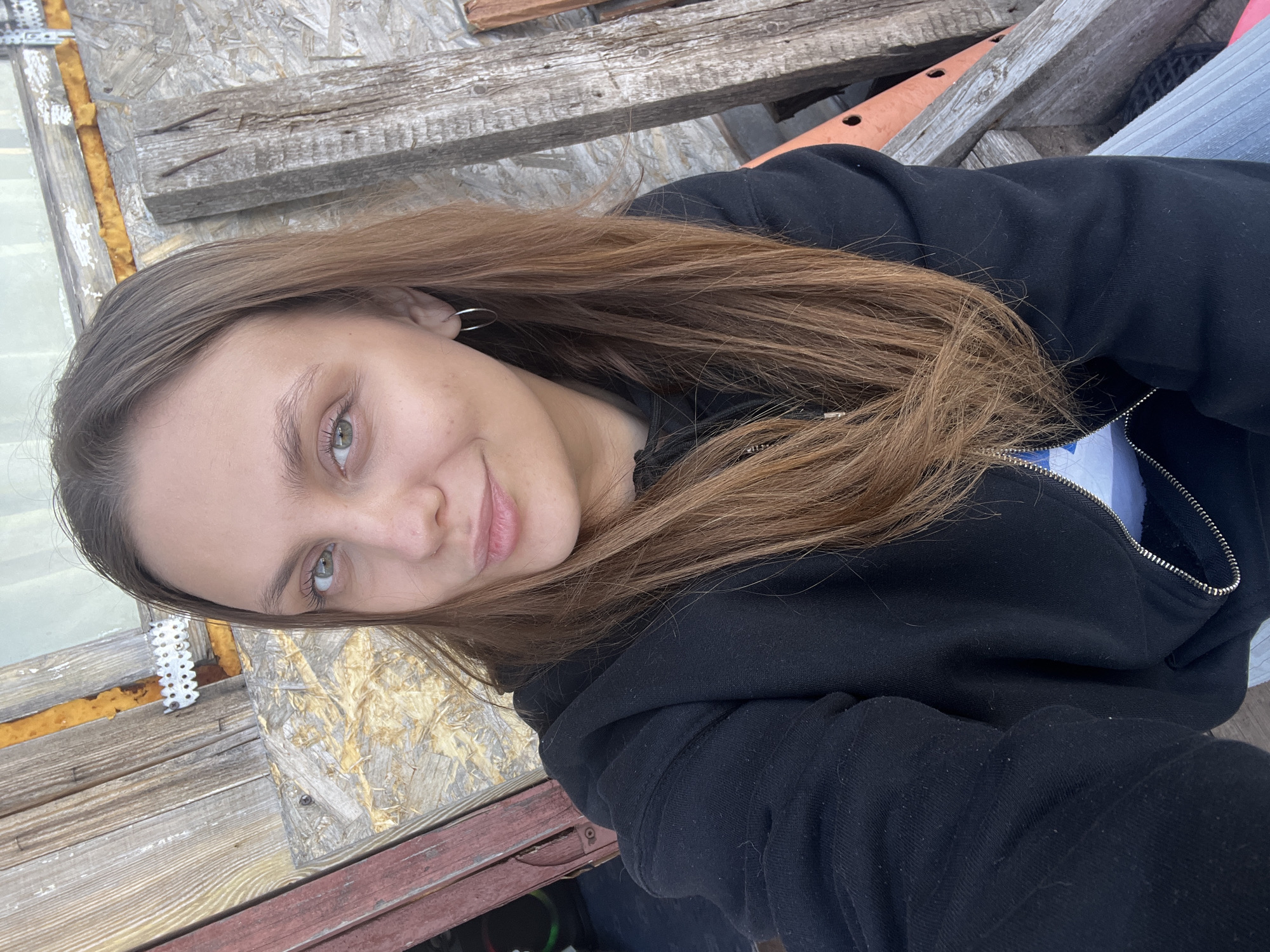 A woman with long brown hair and green eyes is posing for a selfie. She is wearing a black hoodie. The background appears to be a construction site, with exposed wood and building materials. The photo seems to be taken in an outdoor setting. There's no explicit detail indicating the location is Ольховка, Russia. The determination of the location could not be completed to this specification.