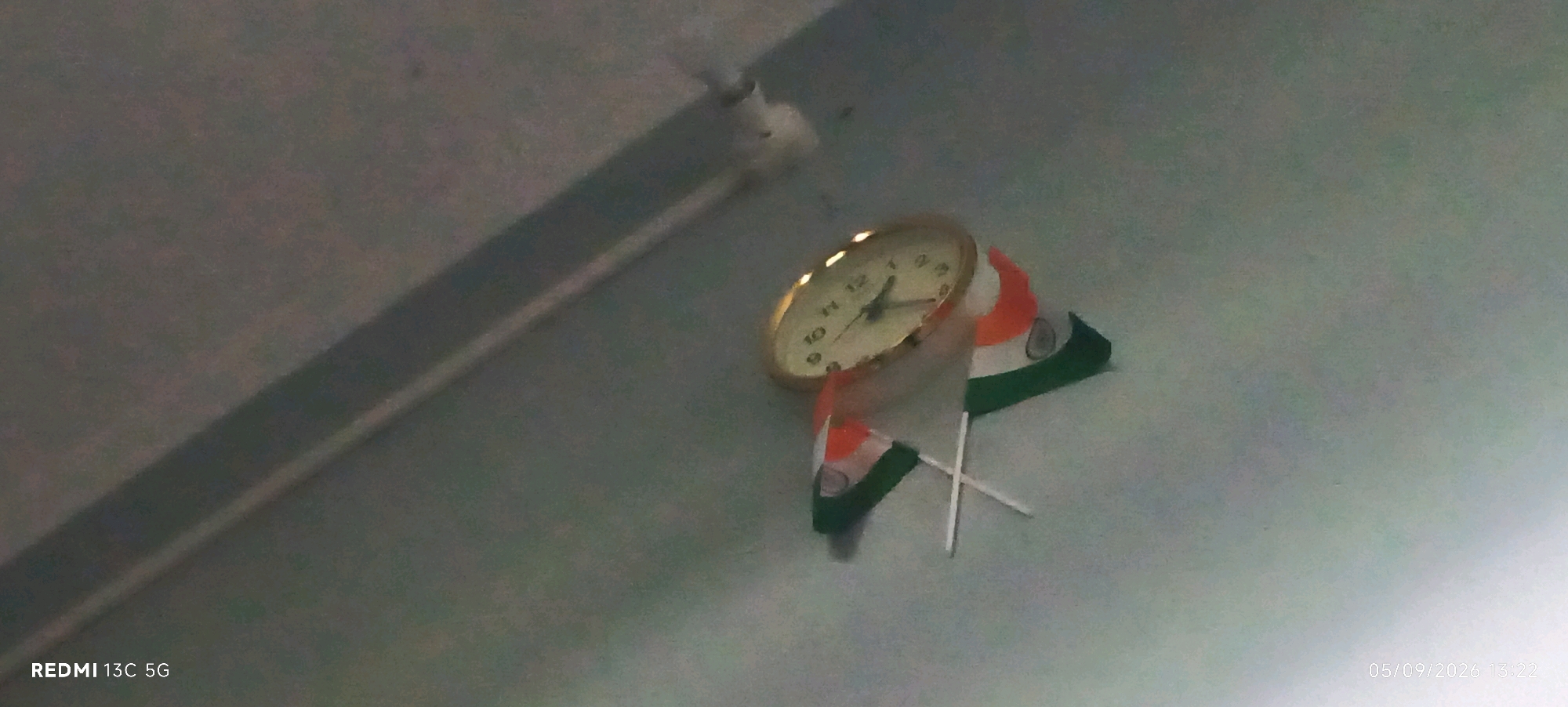 An analog clock with a gold-colored casing is mounted on a wall. Two small Indian flags are attached to the clock, with one flag positioned in front of the clock and the other behind it. The flags are crossed and appear to be made of fabric with a white pole. The wall has a shadow cast upon it, possibly from a light source or an object not fully visible in the frame. The bottom left corner of the image displays the text "REDMI 13C 5G". The bottom right corner shows a date and time stamp: "05/09/2026 13:22". The overall scene suggests an indoor setting, likely a room or an office, in India.