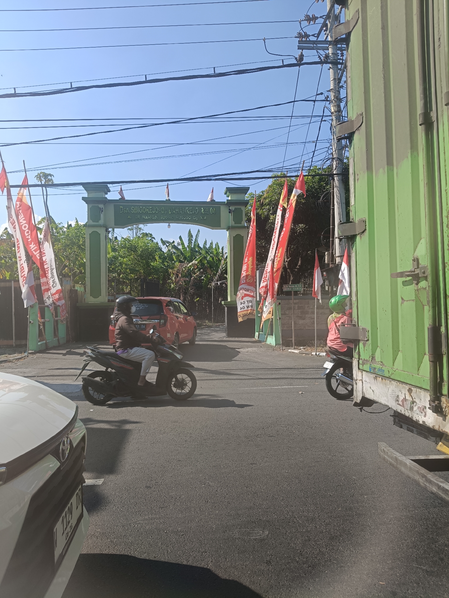The image depicts a street scene in Dahanrejo, Indonesia, likely near an entrance to a residential area or complex. A green archway with text reading "Dsn. Singorejo-Ds. Dahanrejo R/W. 04 Jl. DR. Wahidin Sudiro Husodo Gg. X.4" stands prominently, suggesting a local administrative boundary. Several red and white flags, some with text, are displayed along the roadside and attached to poles.
In the foreground, a white car is partially visible on the left, with its license plate showing "W 1129 DR". To the right of the car, a dark-colored motorcycle is ridden by a person wearing a brown jacket and white pants, with a black helmet. They are moving away from the viewer. Behind the motorcycle, a red car is visible, also in motion.
Further to the right, a large, faded green shipping container is parked, taking up a significant portion of the frame. A person wearing a red jacket and a green helmet is seated on a motorcycle, facing away from the camera, next to the shipping container. The environment is characterized by a clear blue sky, electrical wires strung overhead, and lush green trees and banana plants visible beyond the archway. The road appears to be paved asphalt.