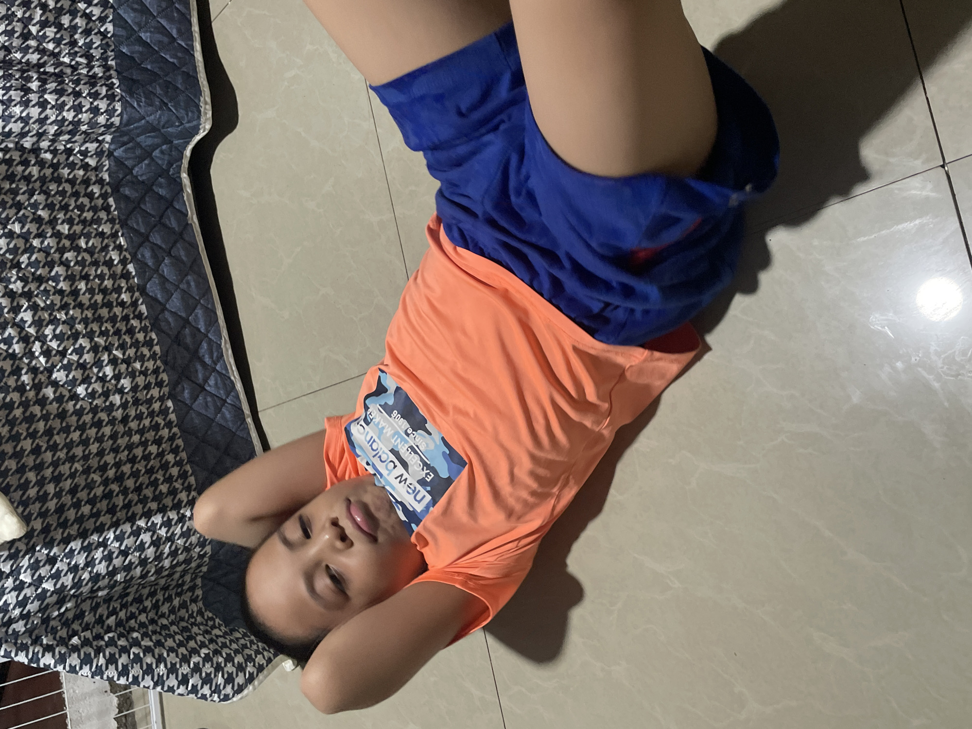 A young child lies on their back on a tiled floor, with their head resting on a patterned mat. The child is wearing an orange t-shirt with a graphic design and blue shorts. The t-shirt has text that reads "New Balance" and "EXCELLENT MAKER SINCE 1906". The child's hands are behind their head, and their legs are bent. The lighting suggests an indoor setting, possibly during the day, as there is a bright light source casting a shadow on the floor. The child's facial expression is relaxed and playful. The patterned mat has a houndstooth design in black and white with a dark blue border. The tiled floor appears to be light-colored and has a marble-like pattern. The scene is likely in Davao City, Philippines, based on the location context provided.