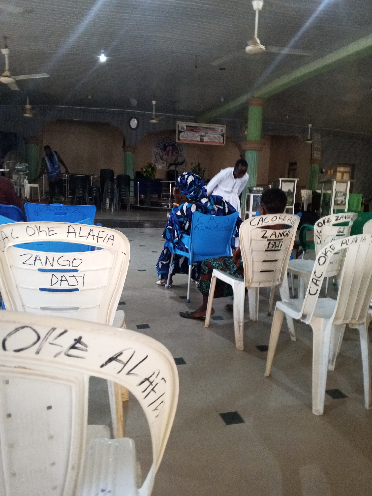 The image shows an indoor space, possibly a church or meeting hall, in Zango-ndaji, Nigeria. Multiple white and blue plastic chairs are arranged in rows, with writing on the backs that include the phrases "OKE ALAFIA ZANGO" and "DAJI". Some individuals are seated on the chairs, including women wearing colorful patterned clothing and head coverings. A man wearing a white shirt appears to be attending to one of the seated women. There are other people standing or moving in the background. The room has ceiling fans, pillars, and framed items on the walls.
