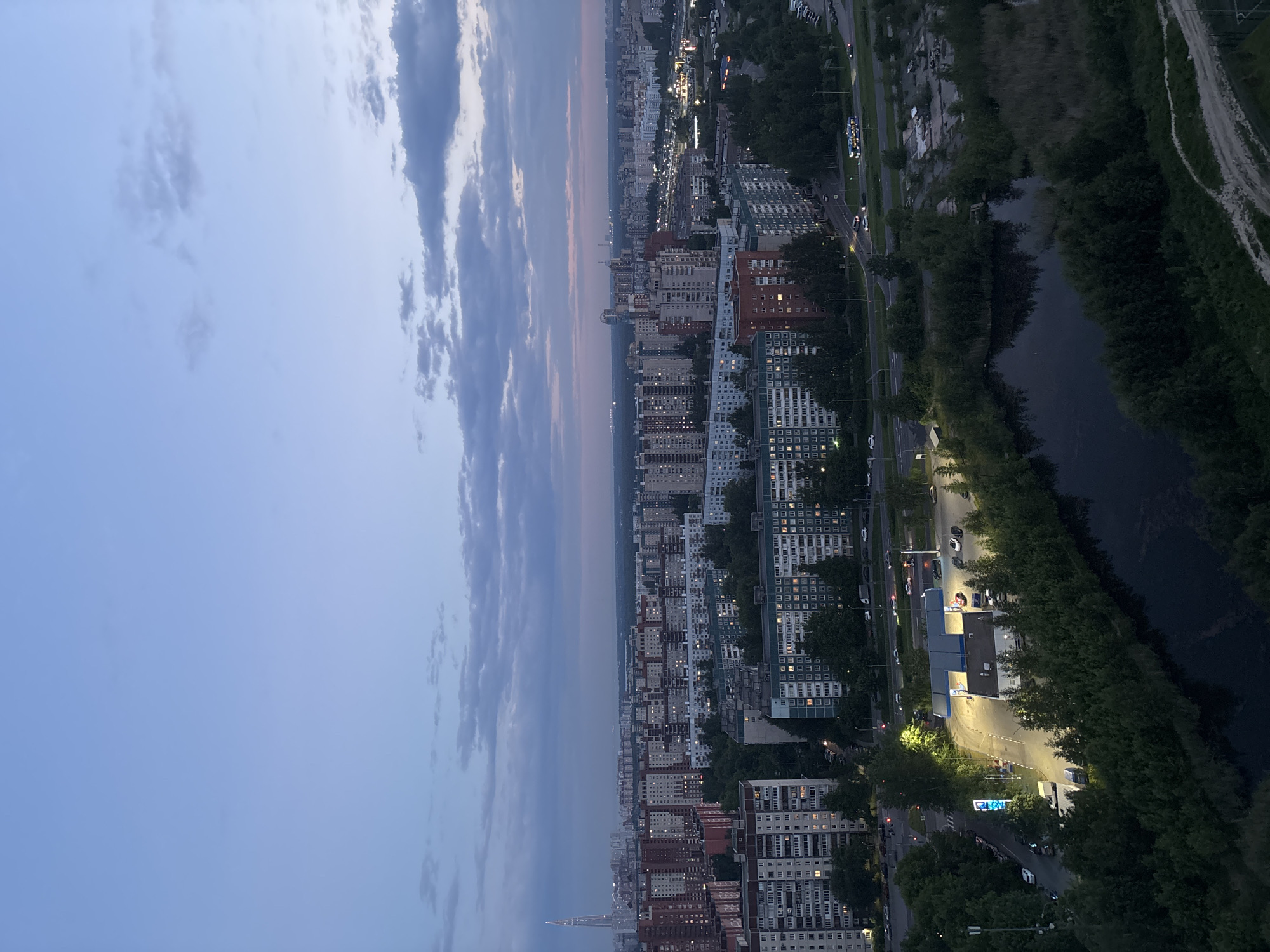 This high-angle, vertical photograph captures an urban landscape at dusk or twilight, looking out over a city transitioning from day to night. The scene is dominated by numerous residential buildings, characterized by their blocky architecture and glowing windows, indicating inhabitants. A significant portion of the image is dedicated to the sky, which exhibits a soft gradient from light blue at the top to a muted pinkish-orange near the horizon, streaked with elongated, dark grey clouds.
In the midground, a densely packed city stretches towards the horizon, illuminated by countless streetlights and building lights that dot the urban fabric. A wide road or highway runs horizontally across the lower right portion of the image, bustling with traffic, as indicated by the streaks of light from vehicles. Adjacent to this road is a dark body of water, possibly a river or canal, lined with dense green trees and vegetation, creating a contrast with the concrete jungle.
A notable landmark, the Lakhta Center skyscraper, is distinctly visible in the distant left, piercing the horizon with its unique, tapering design. Closer to the foreground on the right, there's a brightly lit gas station with cars parked around it, serving as a focal point of activity on the road. The artificial illumination from streetlights and commercial signs casts a warm glow on the immediate surroundings. Although specific text is largely unreadable due to the distance and resolution, the bright blue sign on the gas station appears to be a commercial logo or name. The overall atmosphere is one of a busy city winding down for the evening, viewed from a high vantage point.