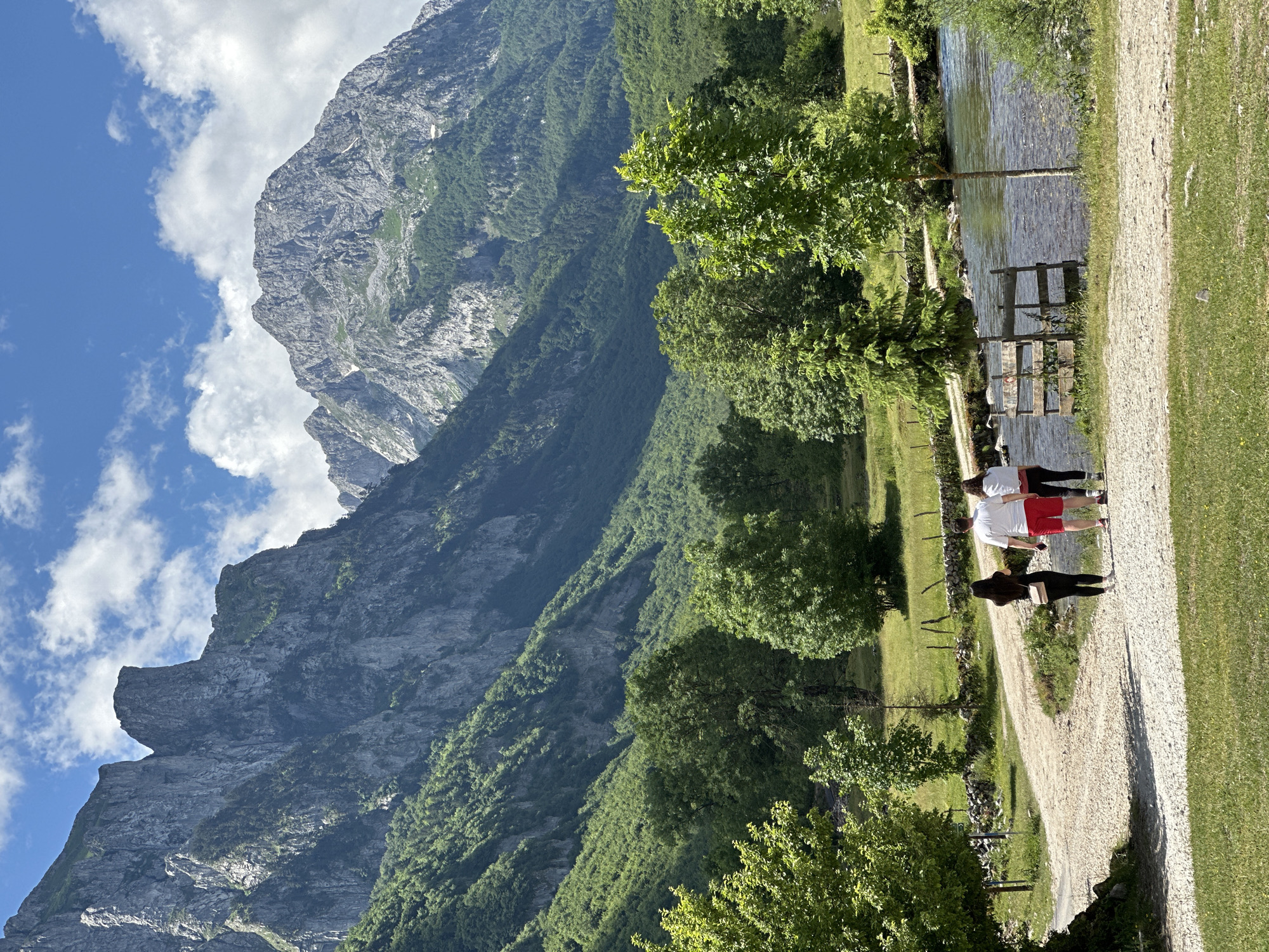 The image depicts a scenic mountain landscape in the vicinity of Gusinje, Montenegro, on a bright, partly cloudy day. In the foreground, three individuals, two women and one man, are walking away from the camera on a light-colored gravel path. The woman on the left wears a black top and pants, carrying a beige purse. The man in the middle is dressed in a white t-shirt and red shorts. The woman on the right wears a white top and black pants.
To the right of the path, a clear, shallow river flows. A rustic wooden fence and a small tree stand near the riverbank. The middle ground features a grassy field interspersed with various green trees of different sizes and densities. Beyond the field, a stone wall and a wooden fence run horizontally.
The background is dominated by majestic, rugged mountains. The slopes of the mountains are covered in dense green vegetation, with visible rock formations and cliffs. The sky above is blue with scattered white cumulus clouds. The overall scene conveys a sense of natural beauty and outdoor recreation.