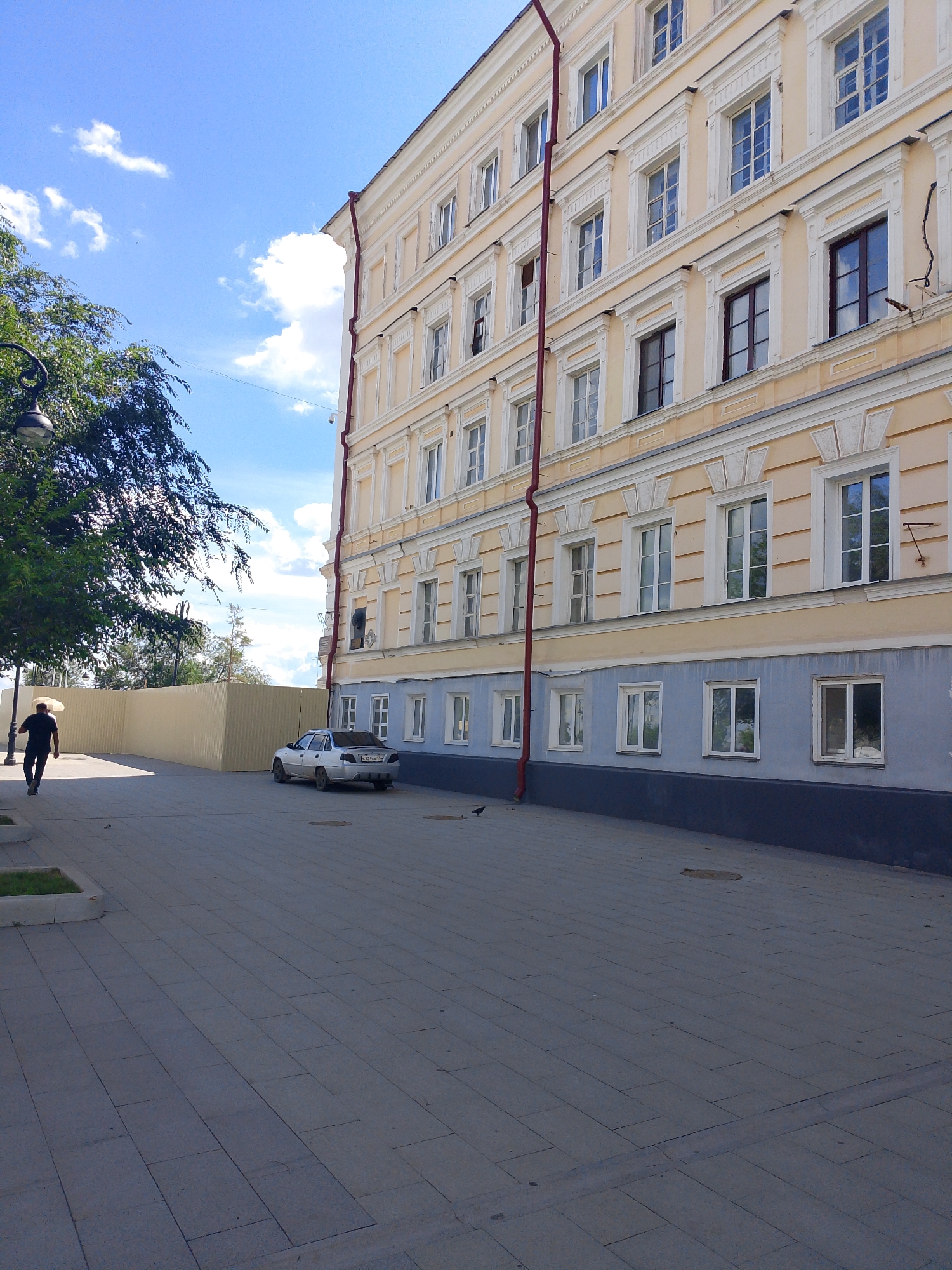The image shows a street scene in Orenburg, Russia, with a large, light yellow building with many windows dominating the right side. A red downspout runs vertically along the building. To the left of the building is a light blue section with more windows. In the foreground, a person in dark clothing walks away from the camera, carrying a light-colored umbrella. A silver car is parked on the street near the building. The ground is paved with large, grey tiles. Lush green trees are visible on the far left, partially obscuring a street lamp. The sky is blue with scattered white clouds. A beige metal fence runs along the side of the street behind the person.