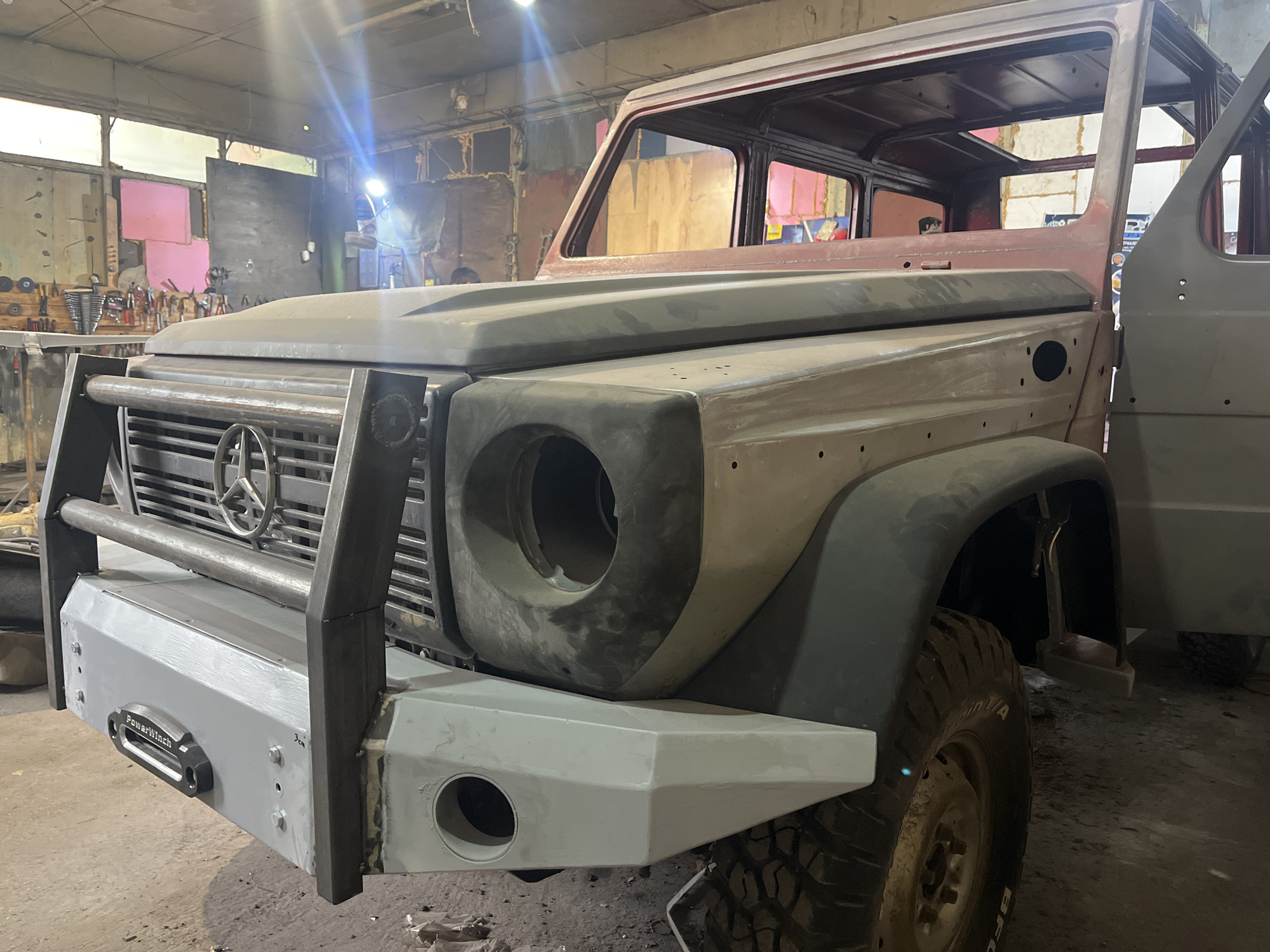 The image shows the front end of a Mercedes-Benz G-Class vehicle undergoing restoration or modification in a workshop setting. The location is likely in Pleven, Bulgaria based on the prompt. The vehicle is in a partially disassembled state, with a prominent brush guard and a newly painted front bumper. The headlamp housings are empty. The body panels are in various stages of preparation, possibly involving body filler or primer. The workshop environment is visible in the background with tools, equipment, and materials related to automotive repair or fabrication. The lighting is a mix of natural and artificial, creating some glare on the vehicle's surfaces.