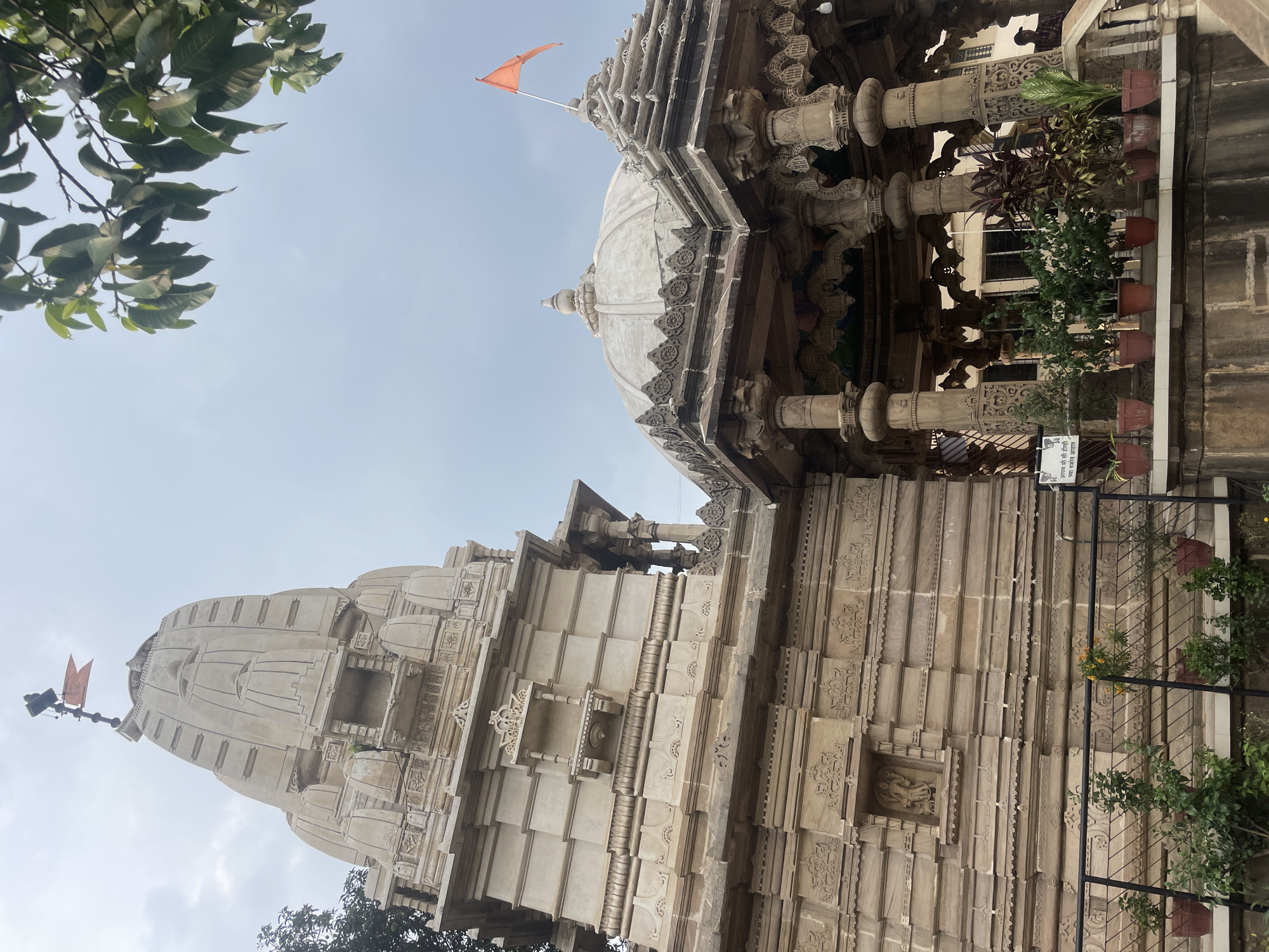 An exterior view displays a stone temple complex under an overcast sky in Kolhapur, India. The primary structure on the left features a tall, light-beige *shikara* (temple tower) adorned with horizontal bands and smaller decorative elements. An orange triangular flag is affixed to a finial atop this *shikara*. The temple wall below includes a carved niche containing a standing figure, possibly a deity.
To the right, an adjoining *mandapa* (hall) is present, characterized by a light-colored, ribbed dome topped with a finial. This *mandapa* is supported by intricately carved stone pillars displaying various motifs. Another smaller orange triangular flag flies from a decorative element on the roofline to the right of the dome.
At ground level, a raised platform is visible, featuring a black metal railing. Numerous potted plants, primarily with green foliage and some yellow flowers, are arranged along this platform. A white sign with Marathi text, partially visible, reads "आपण सीसी टीव्ही" (Aapn CCTV), indicating a CCTV presence. In the background, to the right of the potted plants, a single individual is partially visible, wearing a dark shirt and a cap, near a staircase or elevated area. Lush green foliage from a tree hangs into the frame from the upper right corner.