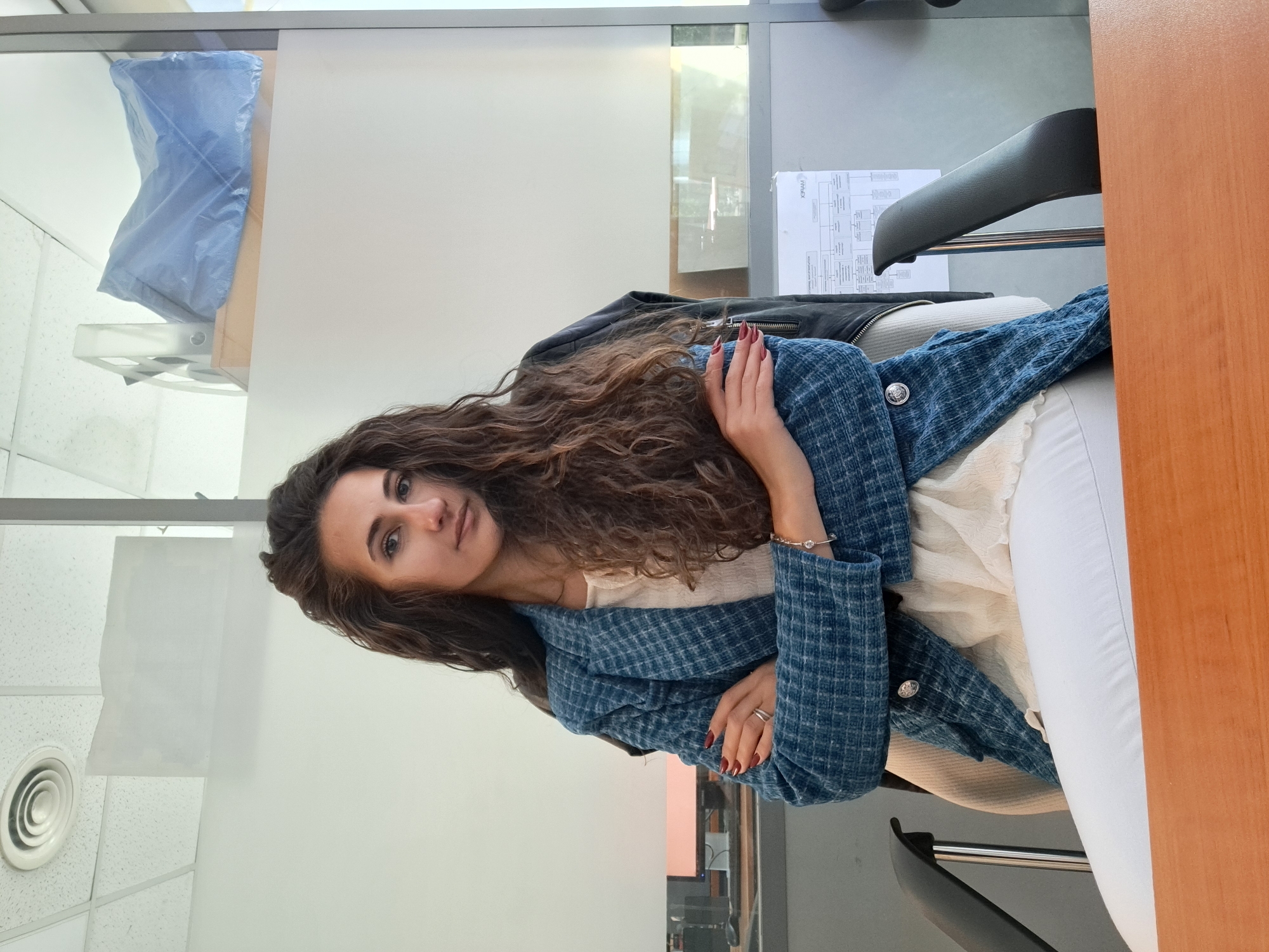 A woman with long, curly brown hair is sitting in a chair in an office setting. She is wearing a white blouse, a blue patterned jacket, and white pants. Her arms are crossed. Behind her, there is a black leather jacket on the chair. The office has a glass wall, and there is a blue object in a clear plastic holder on the wall. There is also a sheet of paper with a diagram or flowchart on it. The location is presumed to be in Sofia, Bulgaria. A desk is visible on the right side of the image.
