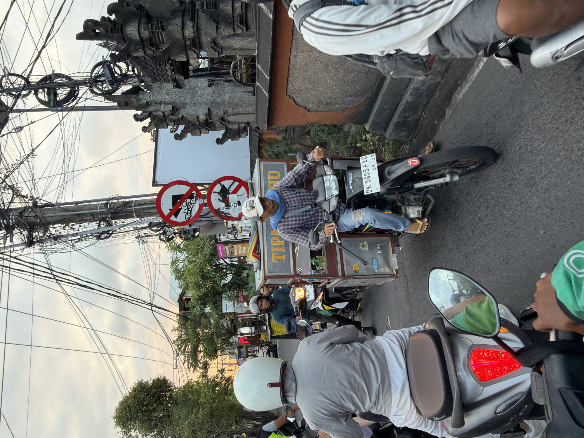 In North Kuta, Indonesia, on a street lined with dense overhead electrical wires and an ornate stone wall, a traffic scene unfolds. Several motorcycles navigate the road, the lead rider in the foreground wearing a white helmet and grey shirt. To the right, a man in a plaid shirt and blue scarf rides a motorcycle pulling a cart. The cart is silver with various items, including what appears to be food or drinks, and signage reading "TIPA." Traffic signs indicating "no left turn" and "no U-turn" are visible. The sky is overcast, suggesting late afternoon or early evening. The road surface is asphalt, and vegetation can be seen on the left side of the street.