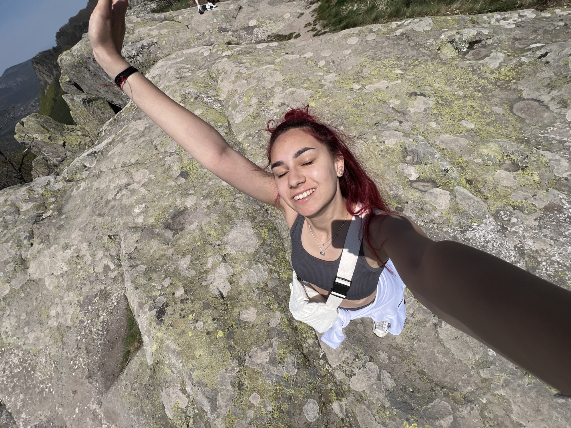 A high-angle selfie captures a young woman standing on a rocky mountain peak at Sini Vrah, Bulgaria, under bright, clear daylight. She holds her right arm outstretched, presumably holding a phone, to take the picture. Her eyes are closed, and she wears a wide, joyful smile, conveying a sense of happiness and relaxation.
The woman has long, reddish-orange hair, pulled back slightly, revealing small earrings. She is dressed in a dark grey ribbed sports bra or crop top, white loose-fitting pants, and white sneakers. A white cross-body bag with a black buckle strap is slung across her body. She wears a silver necklace with a heart-shaped pendant, and a black bracelet adorned with a small red string is visible on her left wrist.
The immediate ground beneath her is a rugged expanse of grey rock, heavily textured and covered in widespread patches of light green and yellow lichen. In the background, other rocky peaks and distant mountains with sparse vegetation are visible under a bright blue sky, confirming a high-altitude outdoor setting. Hints of other people's legs and shoes can be seen in the upper right background, suggesting she is not alone. The strong sunlight casts shadows, indicating a sunny day, likely midday or early afternoon. There is no visible text in the image.