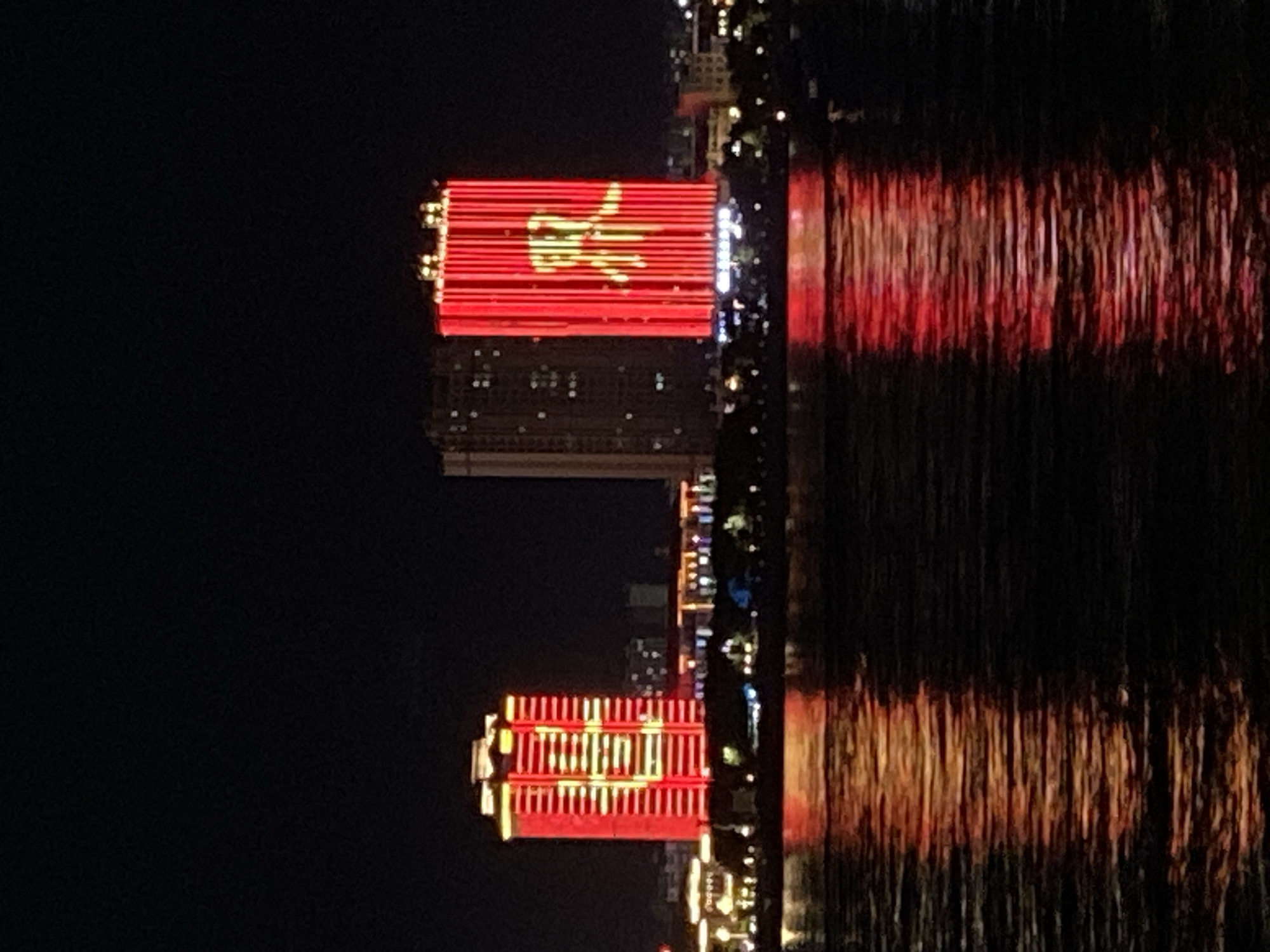 The image shows a nighttime view of the Amur River in Blagoveshchensk, Russia, with illuminated buildings across the river in China. Two prominent buildings are lit with red and yellow lights. The building on the left displays what appears to be Chinese characters or a flag pattern in yellow against a red background. The building on the right features a large, illuminated yellow graphic of a person, possibly a historical or cultural figure, against a red backdrop. The lights from the buildings are reflected in the dark water of the river, creating shimmering patterns of red and yellow. In the foreground, the dark water is choppy, with the reflections of the lights distorted by the movement. The background shows a cityscape with other buildings and lights, also obscured by darkness. The overall scene depicts a modern urban landscape at night, highlighting the illuminated architecture of cities on opposite sides of the river.