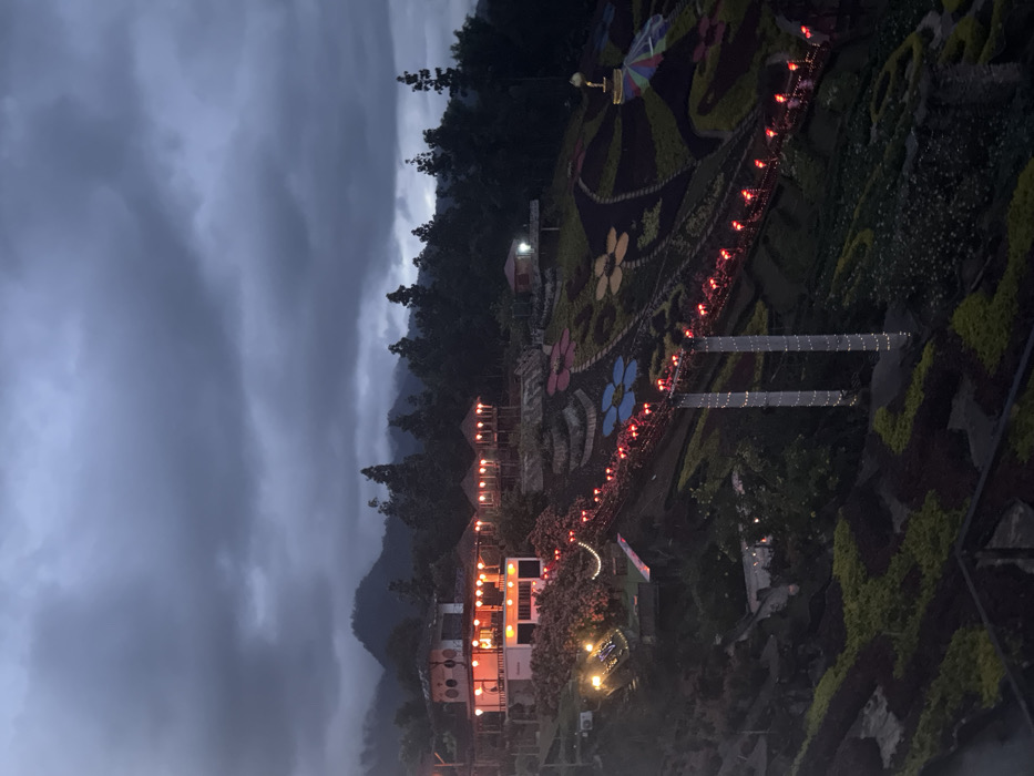 The image depicts a hillside landscape in Ban Mai Saha Sampan, Thailand, at dusk or night under a heavily overcast, dark grey sky. In the background, dark mountains are partially obscured by atmospheric conditions, with dense forest covering their slopes.
On the left side of the frame, a multi-level building with a light-colored facade is illuminated by numerous warm-toned lanterns or string lights, casting an orange glow. Adjacent to this main structure are several elevated, open-sided gazebos with peaked roofs, also lit with warm lights.
The hillside below and to the right of these buildings is extensively landscaped with terraced gardens. These gardens feature large, intricate patterns formed by different colored foliage, including distinct stylized flower designs in blue, yellow, and other hues, along with abstract shapes.
A pedestrian suspension bridge traverses a dip in the terrain, spanning across the landscaped gardens. The bridge's pathway and railings are decorated with numerous bright red lights. Two prominent vertical supports for the bridge are wrapped in white string lights.
Further up the right slope, a smaller, illuminated single-story structure is visible. Within the landscaped garden area to the right, near the bridge, stands a large, multi-colored umbrella-like structure topped with a golden finial or figure. No individuals or specific activities are discernible within the scene.