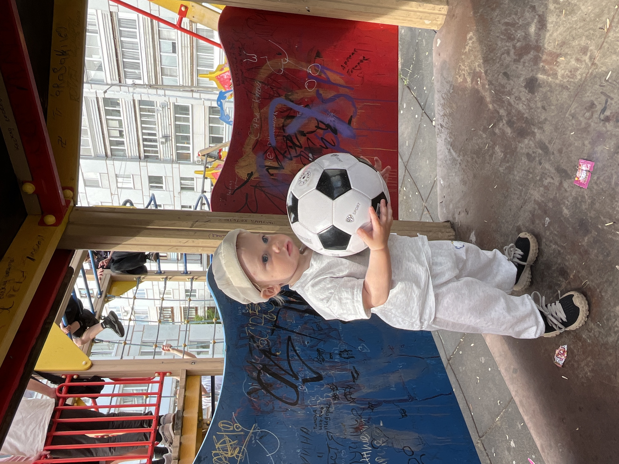 A young child, likely a toddler, is depicted standing on a colorful playground, holding a black and white soccer ball. The child wears a light-colored beanie, a white t-shirt, and white pants, paired with black and white sneakers. The child has light hair and blue eyes, looking directly towards the camera with a neutral expression.
The setting is an outdoor playground, featuring red, yellow, and blue panels, which are heavily covered in various handwritten graffiti and drawings. In the background, tall, modern apartment buildings are visible under bright daylight, suggesting a sunny day in an urban or suburban residential area. Other individuals' legs and arms are partially visible on the playground structure, implying more people are present. The ground is a mix of concrete and dirt, with a pink wrapper lying nearby.
Visible text includes "SPORT" on the soccer ball. On the playground panels, Cyrillic graffiti reads "Сава Кутой" (Sava Kutoy), "TUMDYA," "астя пидарас дуу" (a derogatory phrase), "спыхнуть хDTH TER," "Apreat" (likely a stylized or misspelled "Привет" meaning "Hello"), and "WHA." A swastika-like symbol is also discernible on one of the yellow panels. The scene takes place in Chaikovsky, Russia.