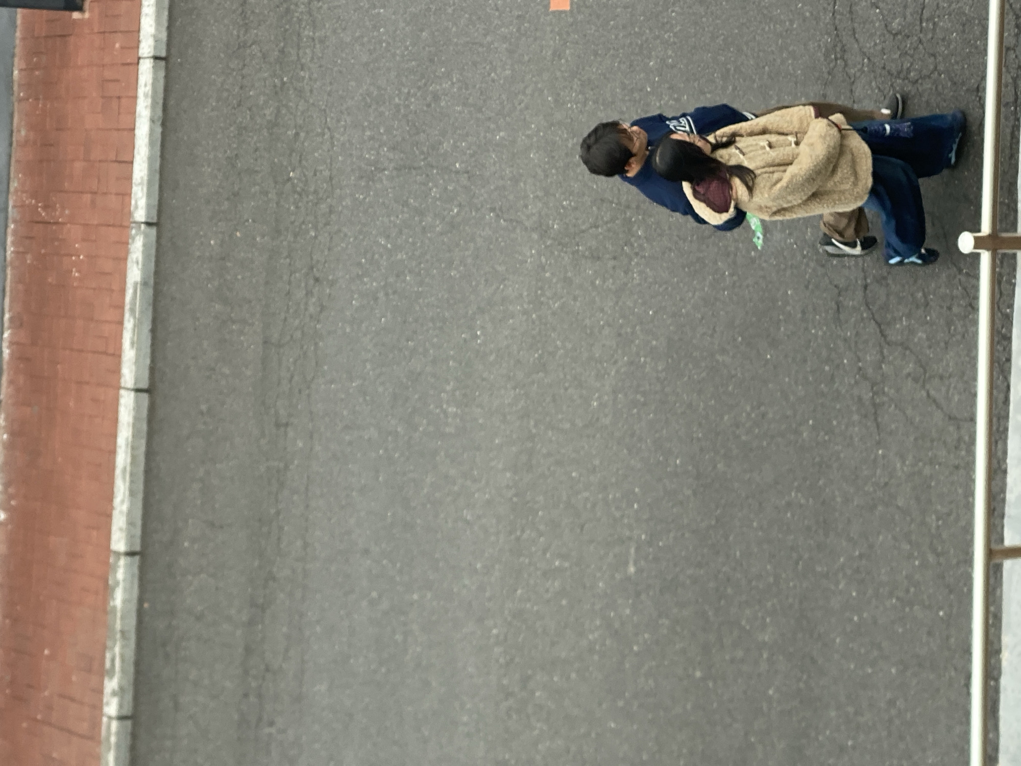 An aerial shot depicts two people standing on a grey asphalt surface. The person on the left wears a dark blue sweatshirt and dark pants. The person on the right wears a tan, hooded jacket, dark pants, and sneakers. The environment is urban, showing a brick wall on the left and a silver railing on the right. The location of Zhongtun, China is unknown.