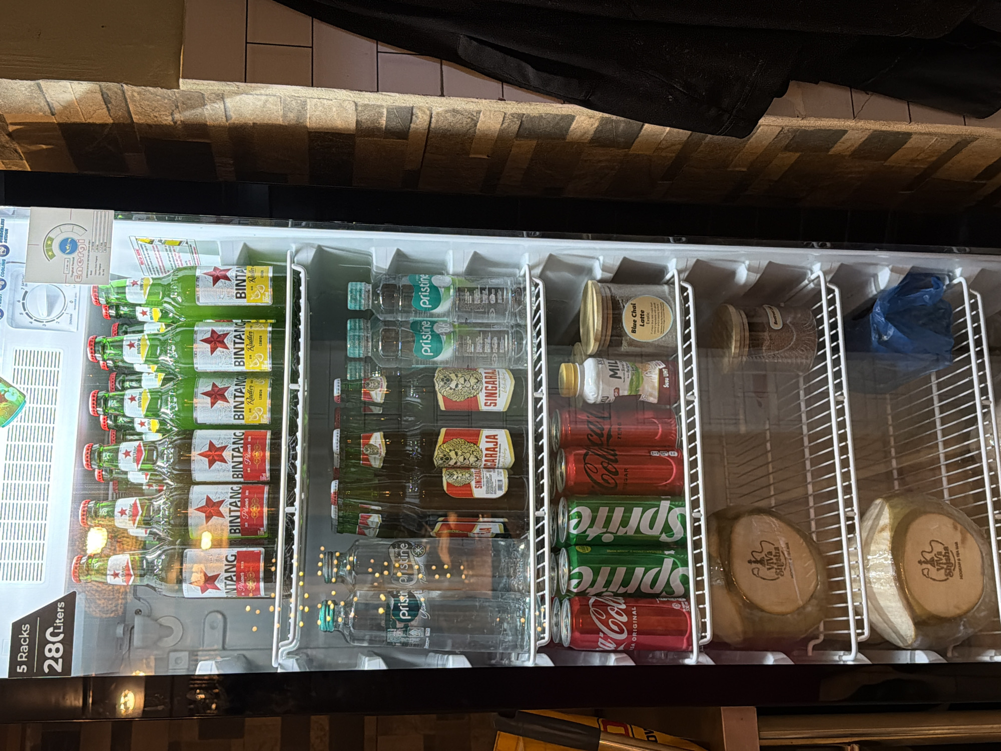The image depicts the interior of a refrigerator in North Kuta, Indonesia, showcasing a variety of beverages. Prominently featured are rows of "Bintang" beer, "Pristine" bottled water, "Singaraja" beer, "Sprite" and "Coca-Cola" cans. Also present are containers labeled "Blue Chai Latte" and "Susu UHT". Two wrapped coconuts with the logo "Viva Shisha" are visible, possibly indicating they are prepared for hookah or tea service.
