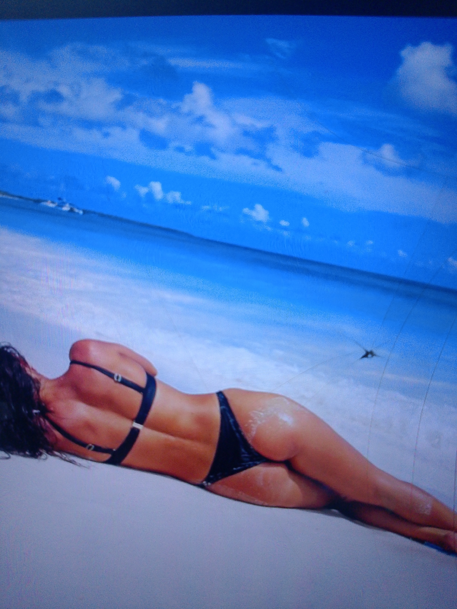 The image depicts a woman in a black bikini lying on a sandy beach next to the ocean. The woman is lying on her stomach, facing away from the camera, with her back and buttocks visible. She has dark hair that is spread out around her head. The beach is light-colored sand and appears to be wet in places. The ocean is a vibrant blue with white waves breaking near the shore. The sky is a bright blue with scattered white clouds. In the distance, there are a few white specks that might be boats. There is also a small, dark object resembling a bird or insect flying in the sky above the ocean. The scene is brightly lit, suggesting it is daytime. There is no indication of the city of Dobra Krynytsia, Ukraine, in this image.