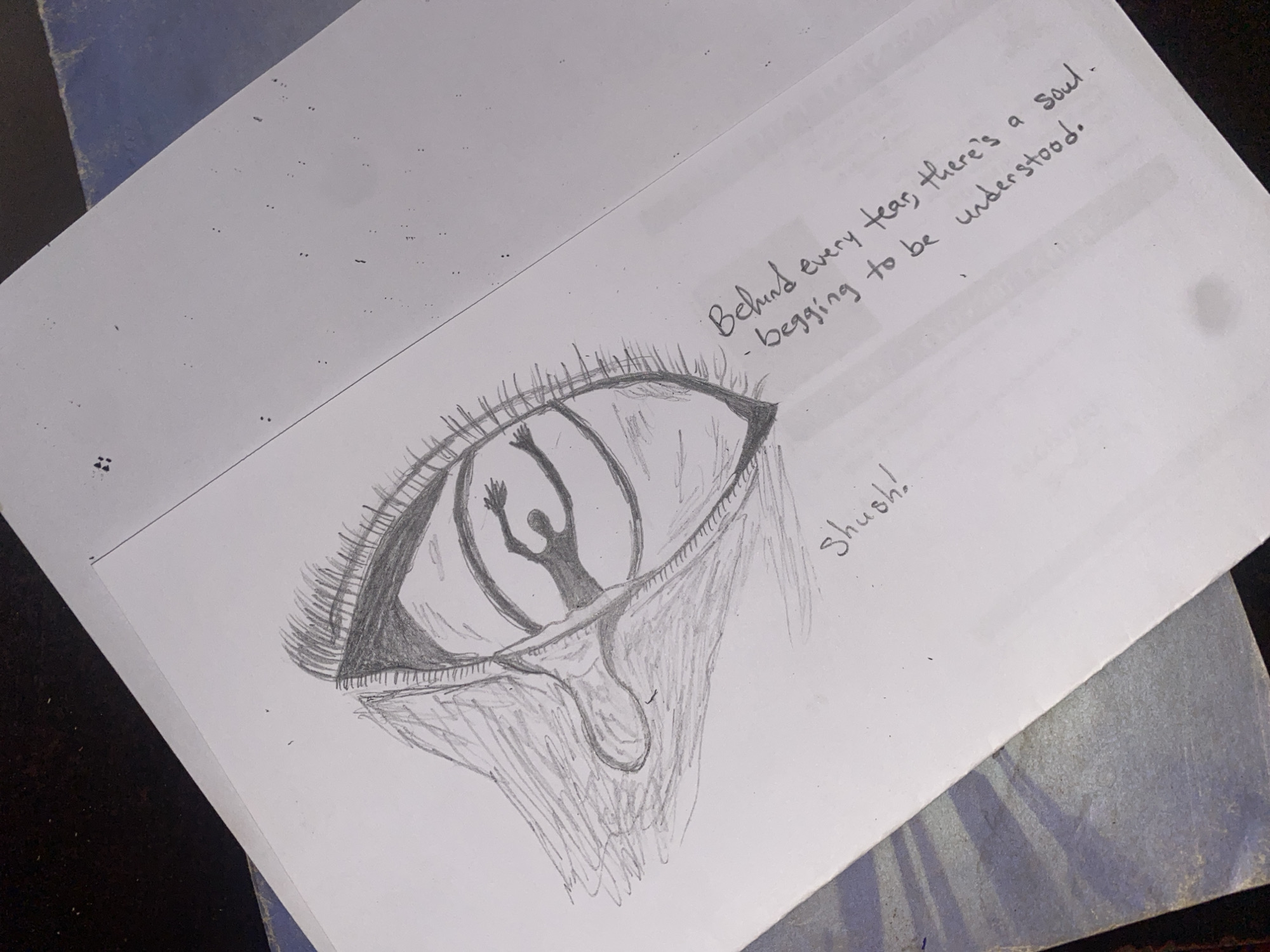 A pencil drawing on a white sheet of paper depicts a large eye crying. Inside the iris, a small figure appears to be struggling with its hands raised. Beneath the eye, the tear is heavily shaded with pencil strokes. Above the eye, handwritten text reads, "Behind every tear, there's a soul - begging to be understood." To the right of the eye, the word "Shush!" is also handwritten. The paper rests on a dark surface with a partially visible blue object beneath it. The scene does not contain elements identifying Rido, Nigeria.