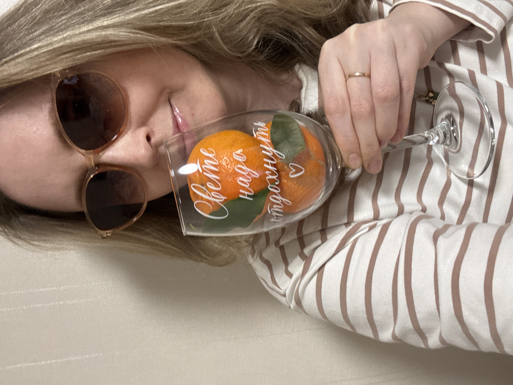 A woman with blond hair and sunglasses holds a wine glass containing two mandarin oranges with green leaves in front of her face. The glass has text written on it in Cyrillic script. The woman is wearing a white shirt with brown stripes. She is wearing a wedding ring on the ring finger of her left hand. The background appears to be a light-colored wall. The location is Chaikovsky, Russia.
