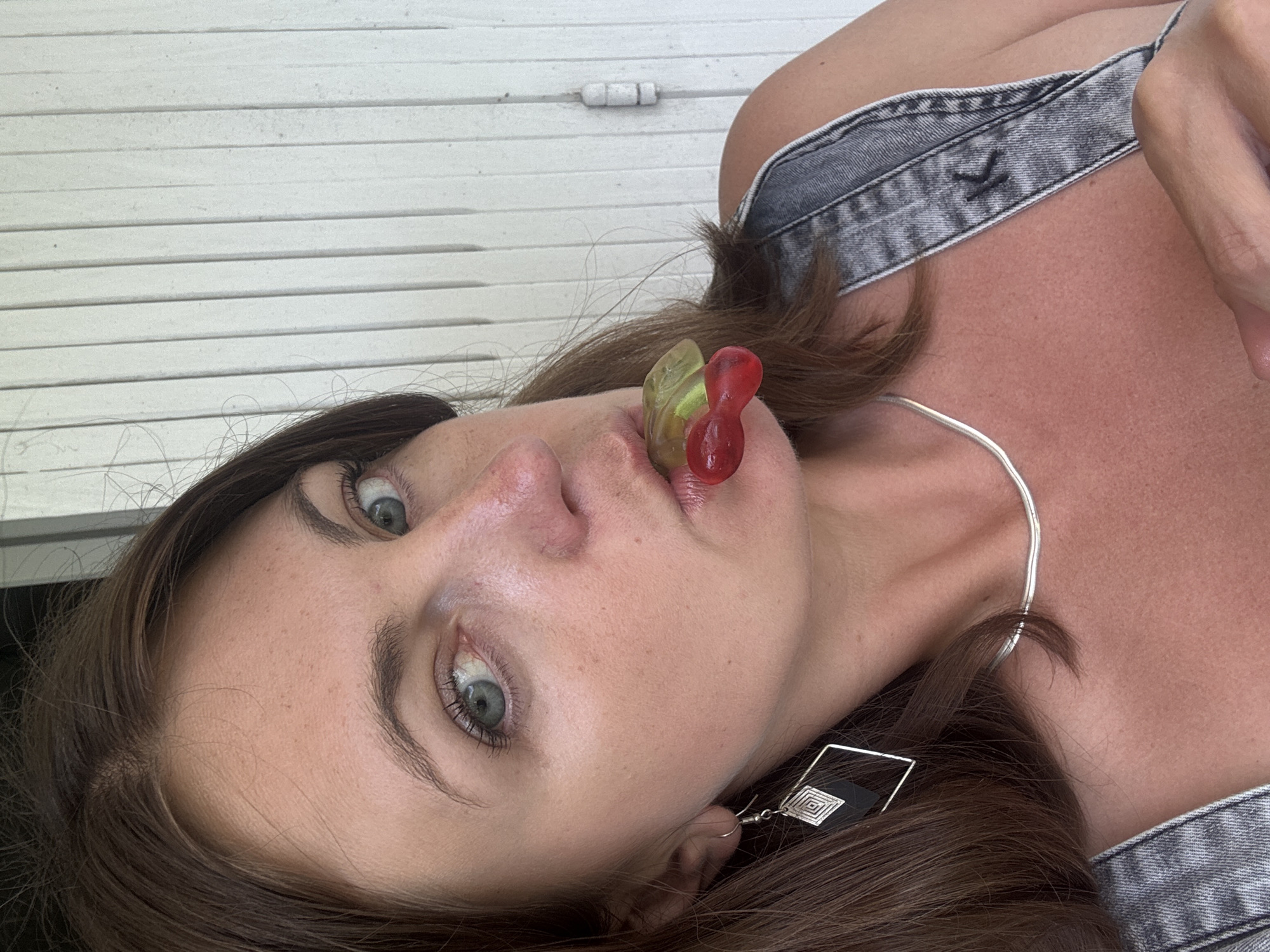 The media file is a close-up, horizontally oriented photograph of a young person's face and upper torso. The subject has brown hair, fair skin with some freckles, and striking blue-green eyes. They are wearing denim overalls and a silver necklace. Two gummy candies, one red and one green, are held between the subject's lips. The subject's head is tilted, and their gaze is directed towards the camera, conveying a playful or curious expression. The background consists of white horizontal slatted blinds, suggesting an indoor setting or a sheltered outdoor area. The lighting is bright, hinting at daytime. There is no visible text in the image. The context provided places the scene in Saint-Claude, France.