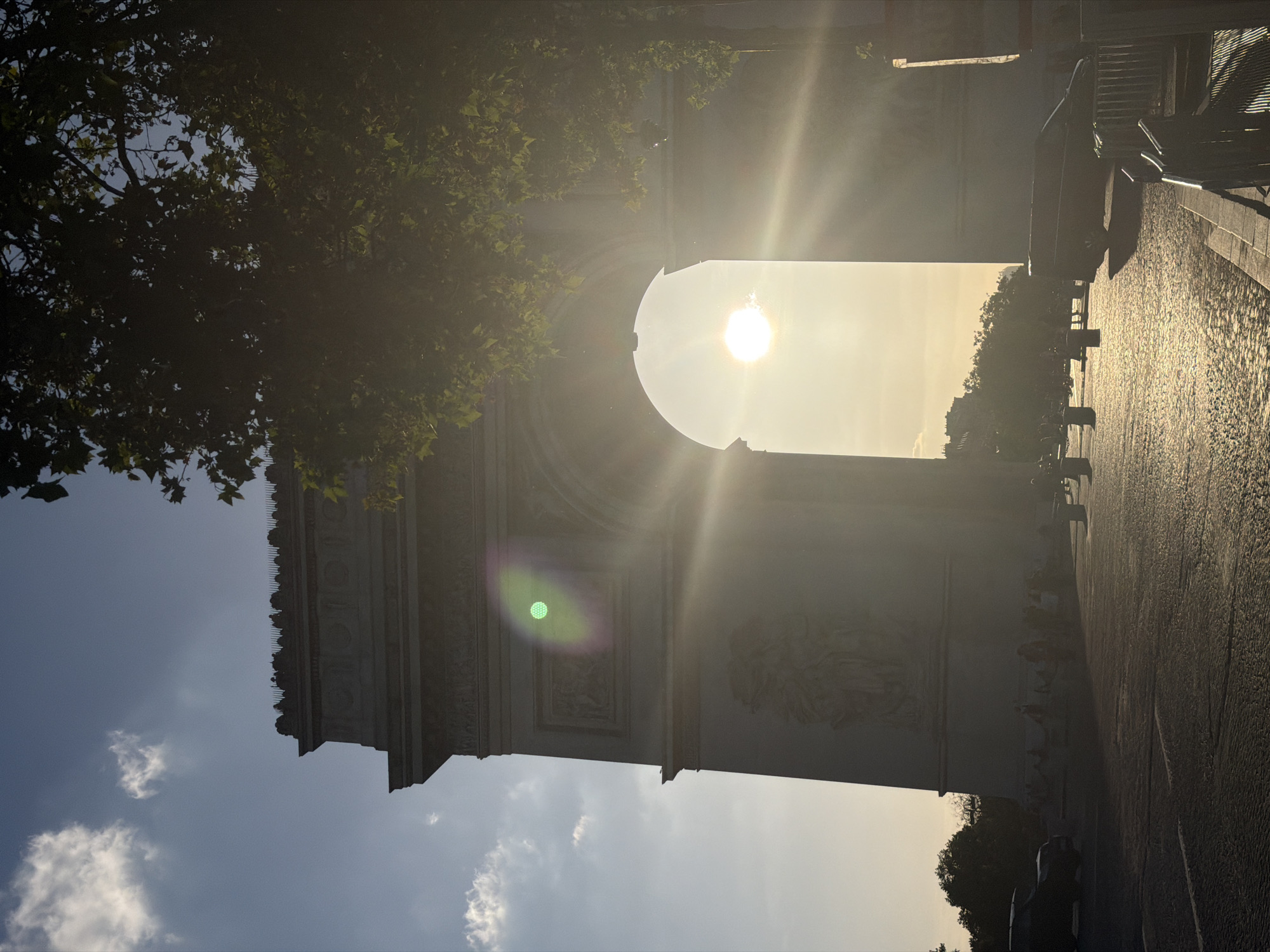 The image shows a view of the Arc de Triomphe in Paris, France, under bright sunlight. The sun is positioned directly in the center of the arch. A tree branch obscures part of the upper left corner. A sidewalk and part of a barrier are visible in the upper right. The sky is visible on either side of the Arc de Triomphe.
