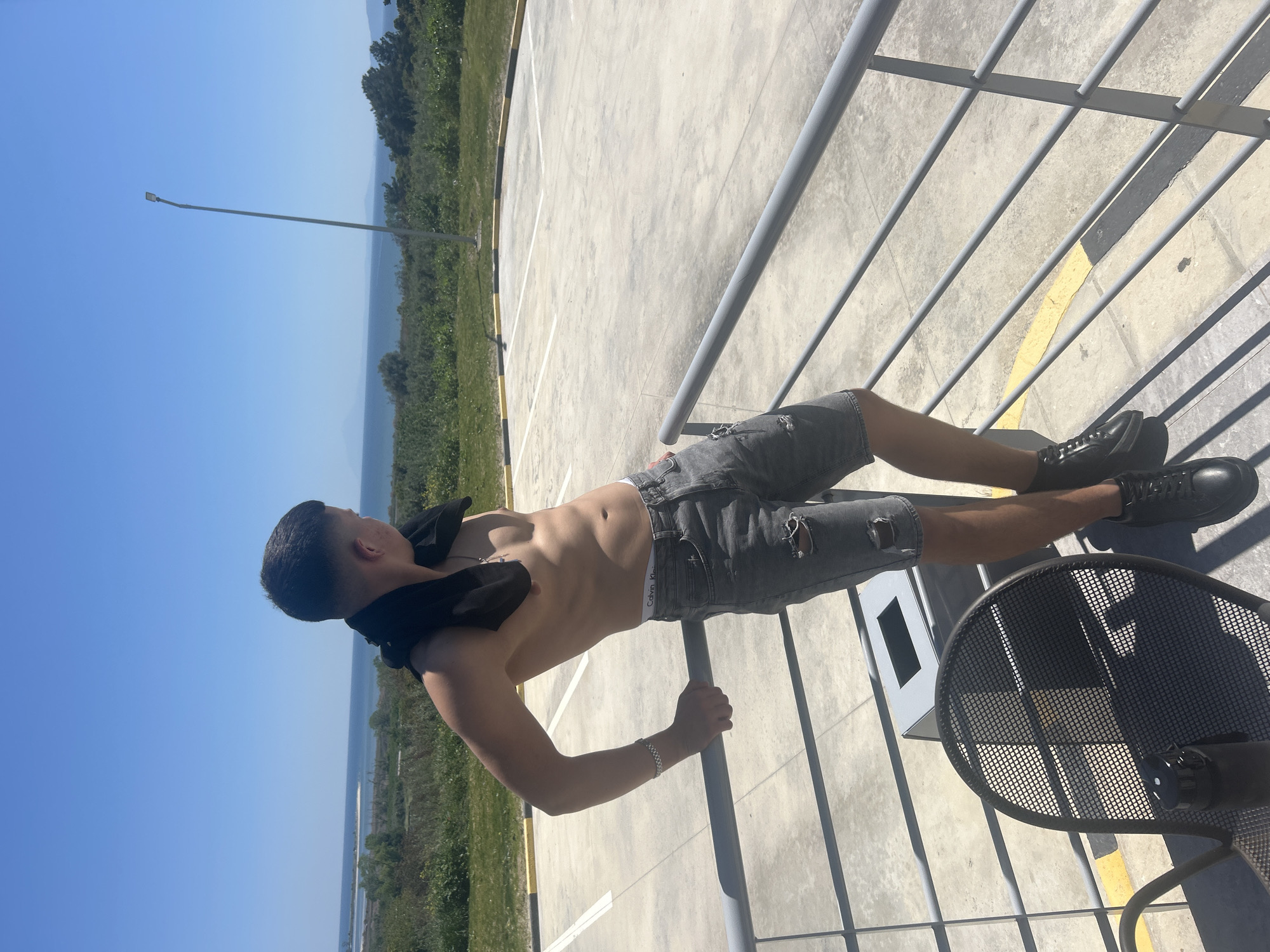 A young man stands shirtless, holding onto a metal railing. He is wearing ripped grey shorts and black shoes. A black garment hangs around his neck. Behind him is a concrete expanse, bordered by trees and a distant coastline under a clear blue sky. A metal table and trash bin are visible near the railing. The location is Νέα Κερδύλια, Greece.