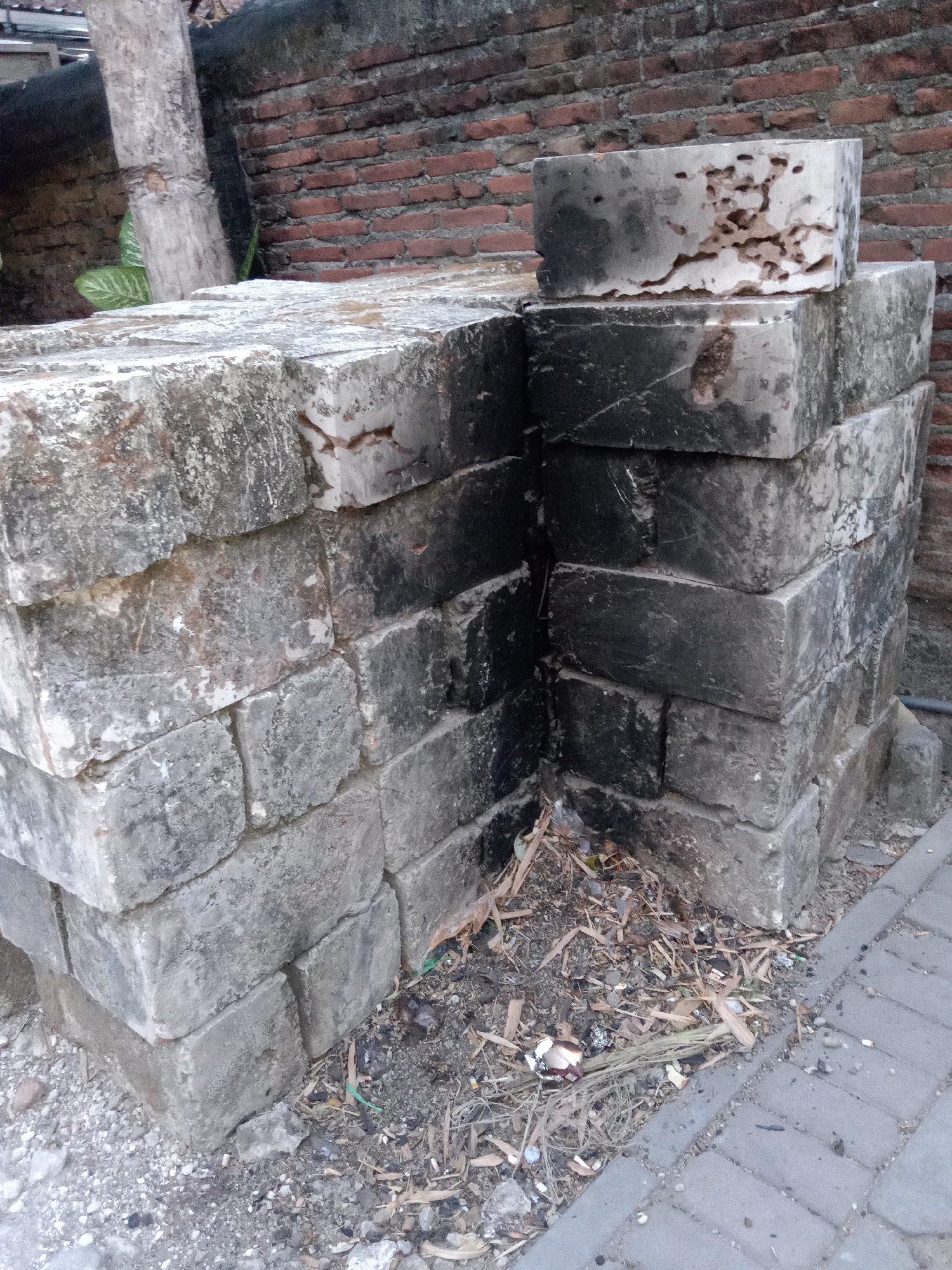 A stack of weathered, gray concrete blocks stands in the foreground, forming a corner structure. The blocks exhibit signs of age and wear, with some showing discoloration, cracks, and pitting. One block at the top appears to have been damaged by insects, with visible tunnels. The stack is partially obscured by a dry, woody plant and debris on the ground, including dried leaves, twigs, and small pebbles. Behind the concrete blocks, a red brick wall is visible, constructed in a horizontal pattern. To the left of the blocks, a rough, textured tree trunk is present, with a green leaf visible near its base. The ground is a mix of dirt and paved interlocking gray pavers. The scene appears to be an outdoor area, possibly a construction site or storage area, located in the city of Jumput, Indonesia. There are no people or animals visible in the image.