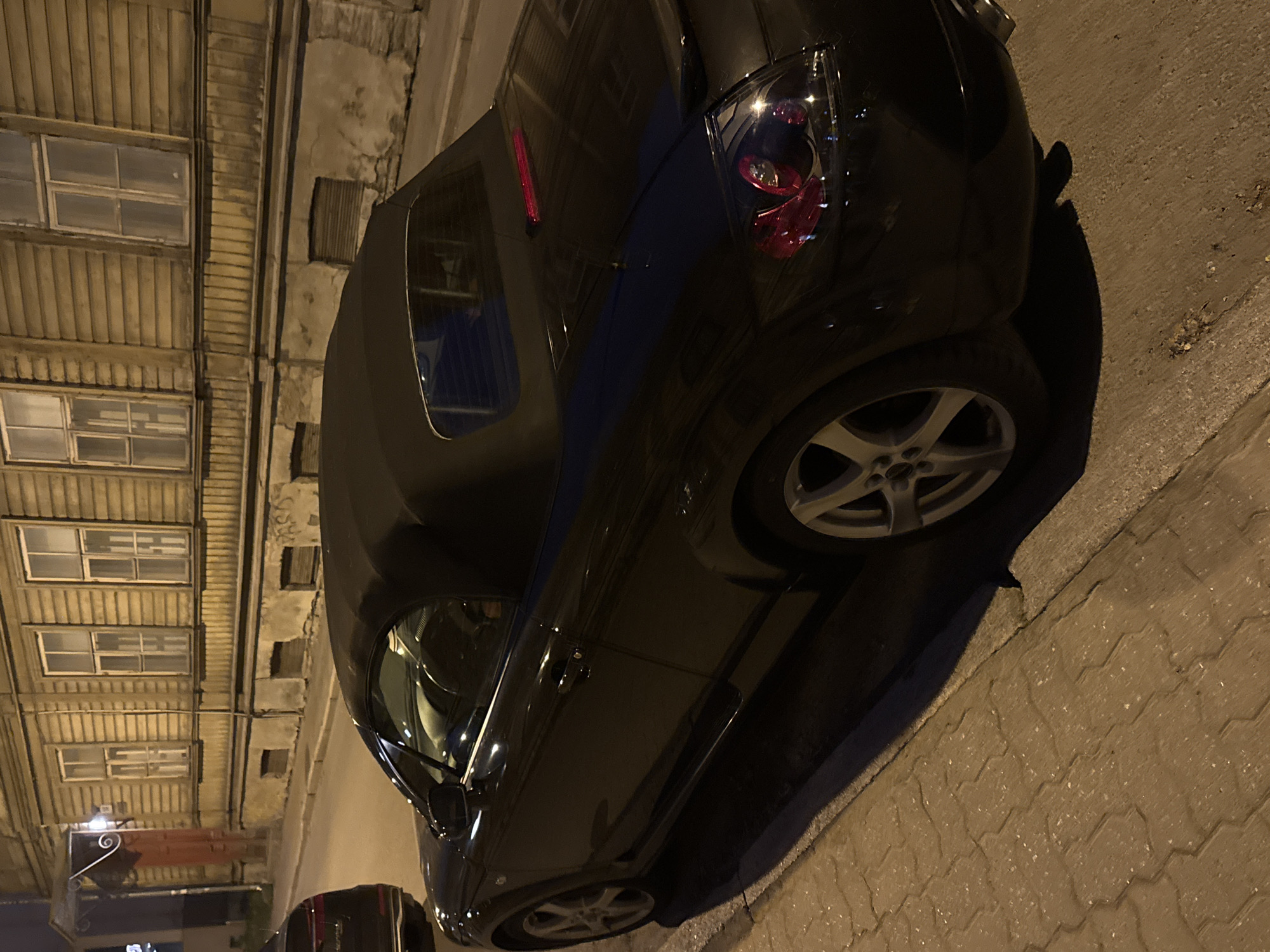 This is a medium shot, taken at night, of a black convertible car parked on the street in Tallinn, Estonia. The car is the primary subject, angled from the rear passenger side, showing its sleek design and a partially visible interior through the open top. The car's taillights are illuminated, casting a red glow.
The car is parked on a cobblestone sidewalk next to a street. In the background, there is a weathered, light-colored building with multiple windows. The building's facade appears to be made of wood planks and has aged stonework at the base. The streetlights illuminate the scene, creating shadows and highlighting the texture of the cobblestones. A portion of another black vehicle is visible in the far left of the frame, suggesting it is also parked on the street. There are no people visible in the image.