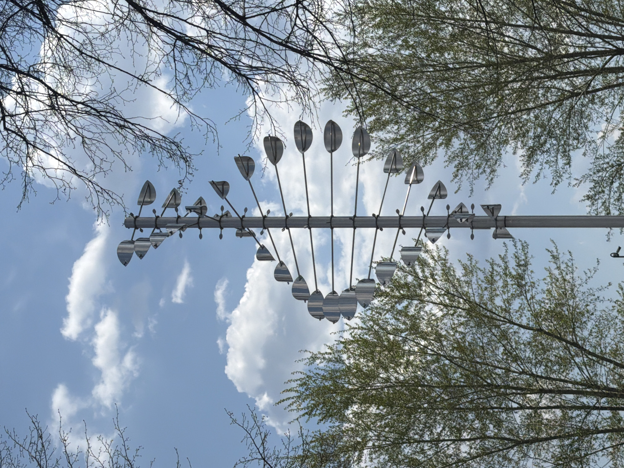 The image depicts a kinetic wind sculpture set against a backdrop of a partly cloudy sky, framed by tree branches. The sculpture consists of a horizontal metal beam with numerous reflective, leaf-shaped elements suspended from it. The leaves are arranged in a symmetrical pattern. Bare tree branches occupy the upper-left corner of the frame, while trees with budding leaves are visible on the right. The sky is blue with scattered white clouds. There is no indication of any people in the scene. Based on the metadata, this image was taken in Votkinsk, Russia.